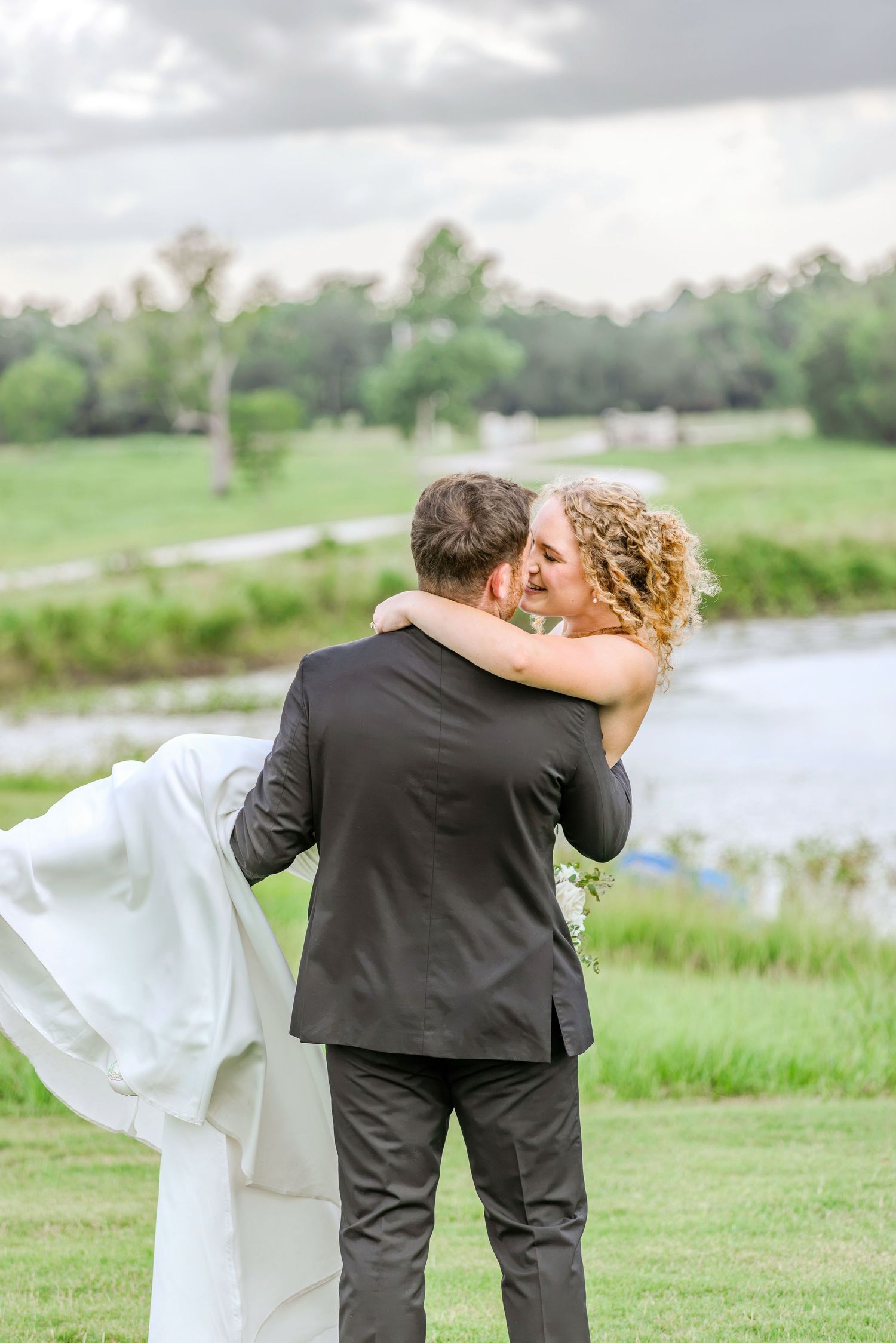 Bride and groom share a romantic kiss outdoors near a pond, with the bride in a flowing white gown on a cloudy day.