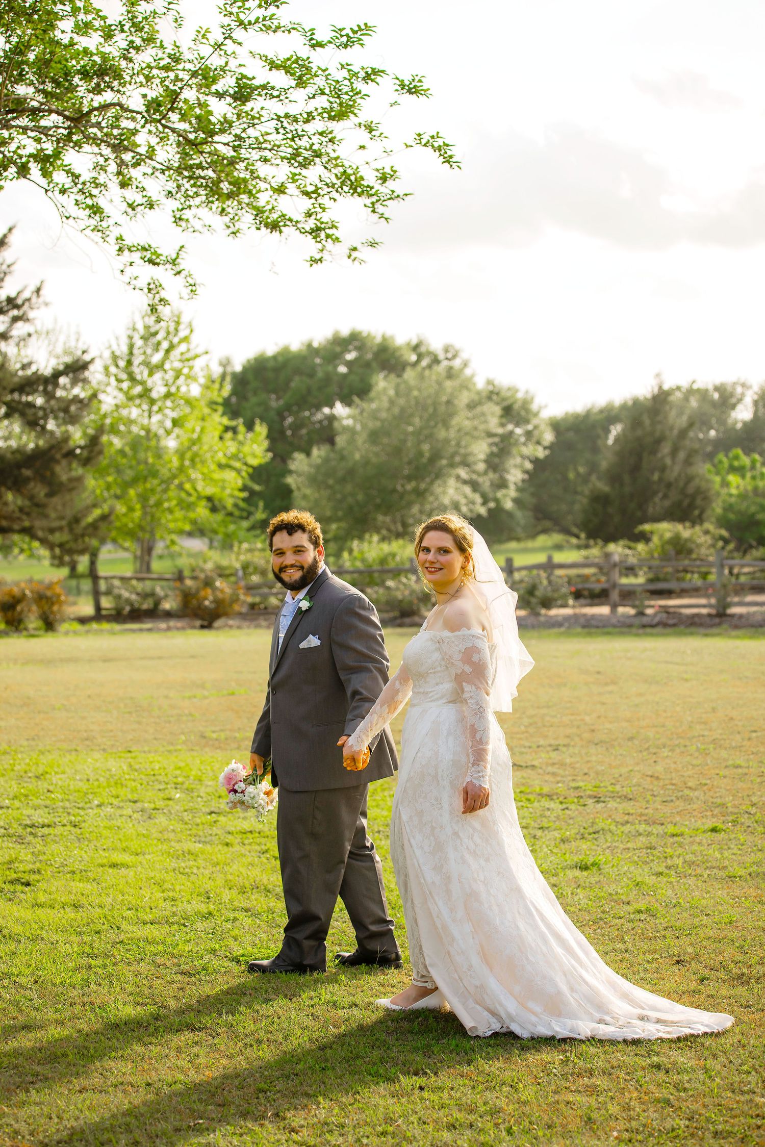 Bride and groom holding hands walking on green lawn in wedding attire, lace dress with veil and gray suit.