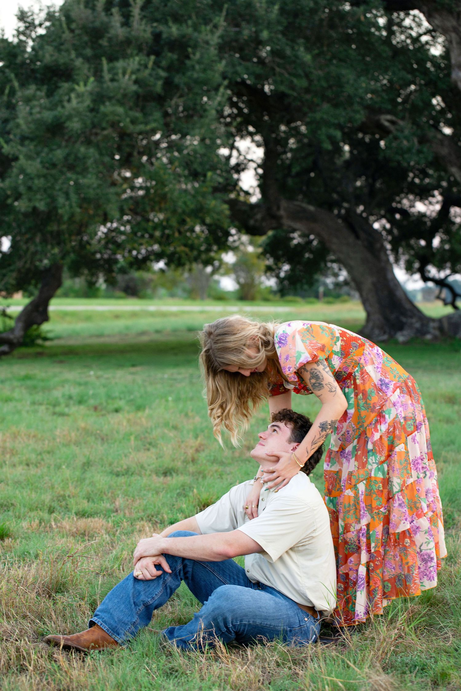 A couple sits together in a grassy field on a sunny day, sharing an intimate moment under the trees.