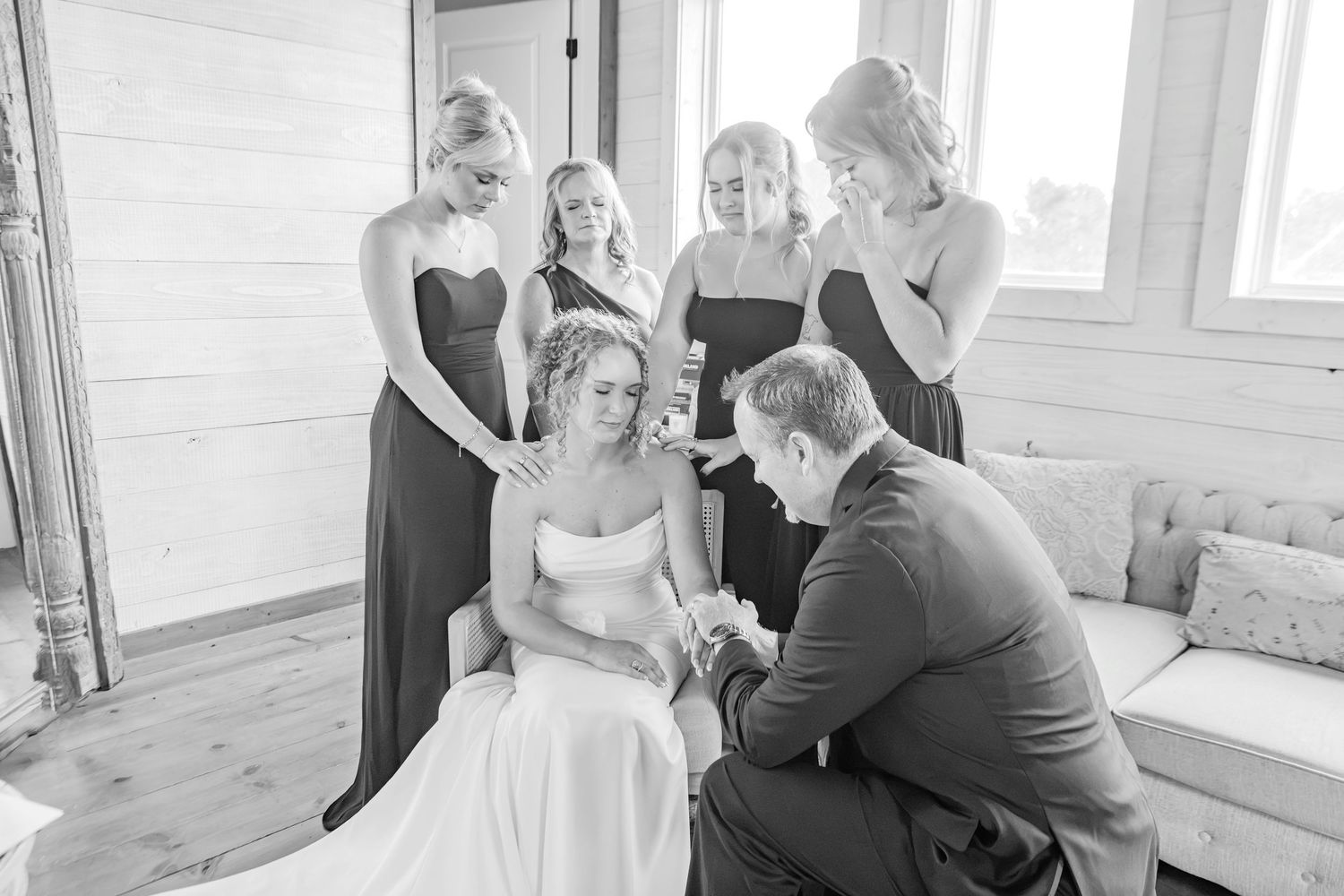 Black and white photo of a bride surrounded by bridesmaids and groom in prayer before the wedding ceremony.