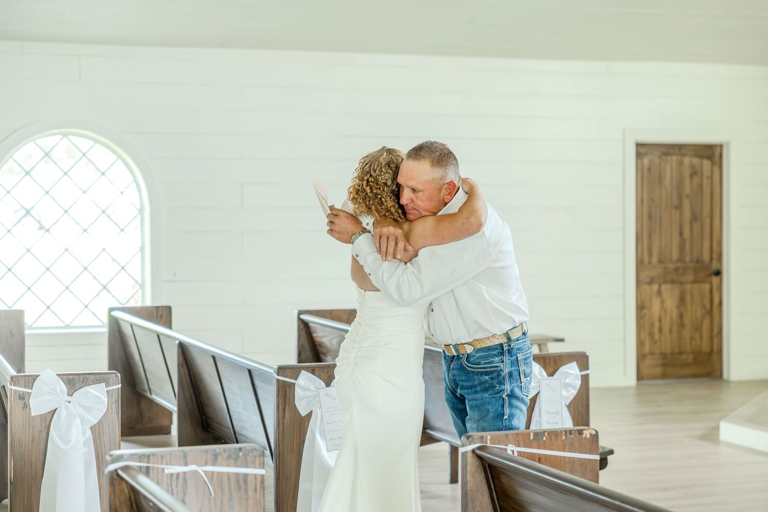 Bride and groom sharing a tender hug in a white chapel with wooden pews decorated with white ribbon bows.
