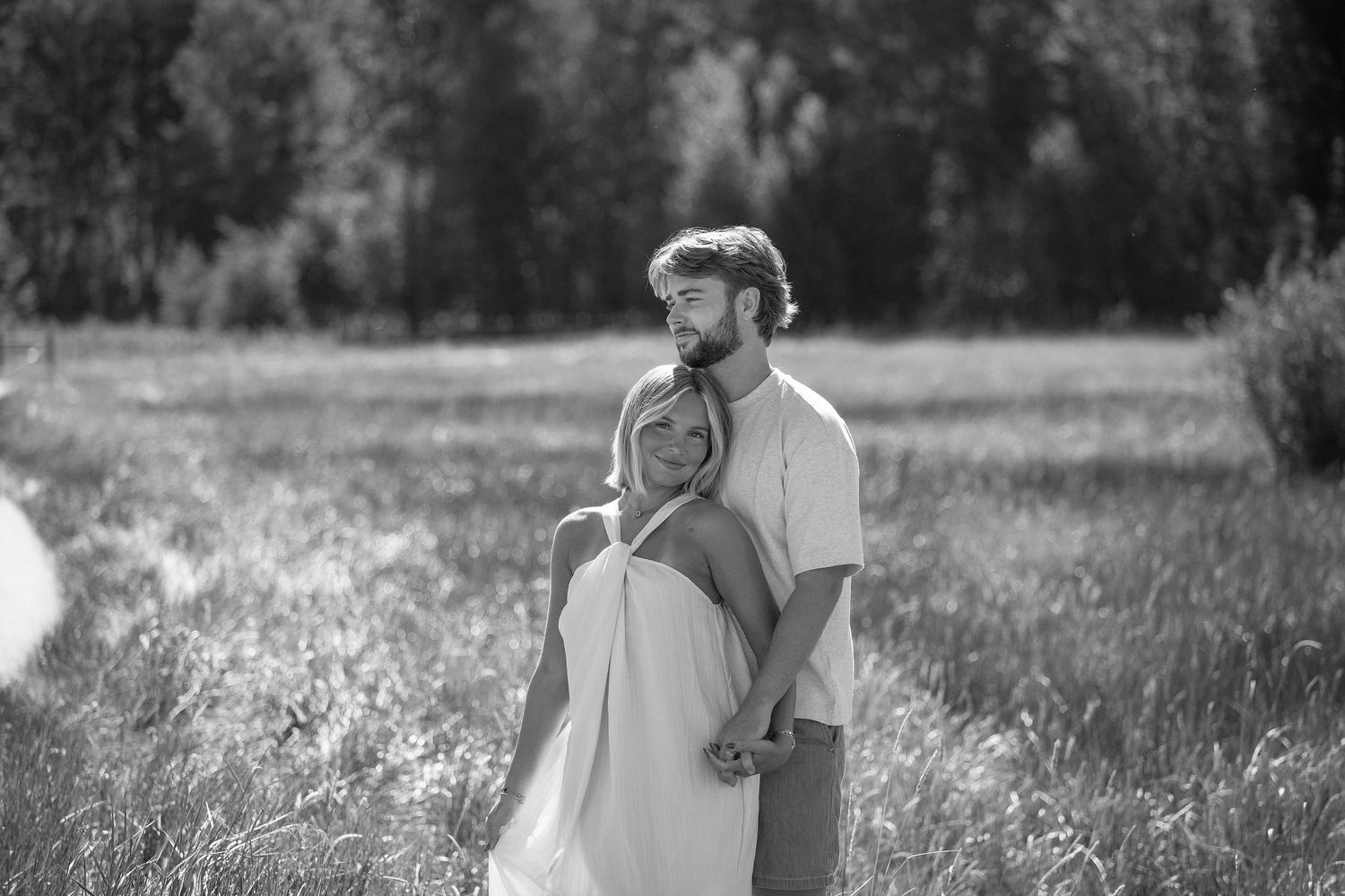 Black and white photo of a couple embracing in a field, man kissing womans head, trees in background.