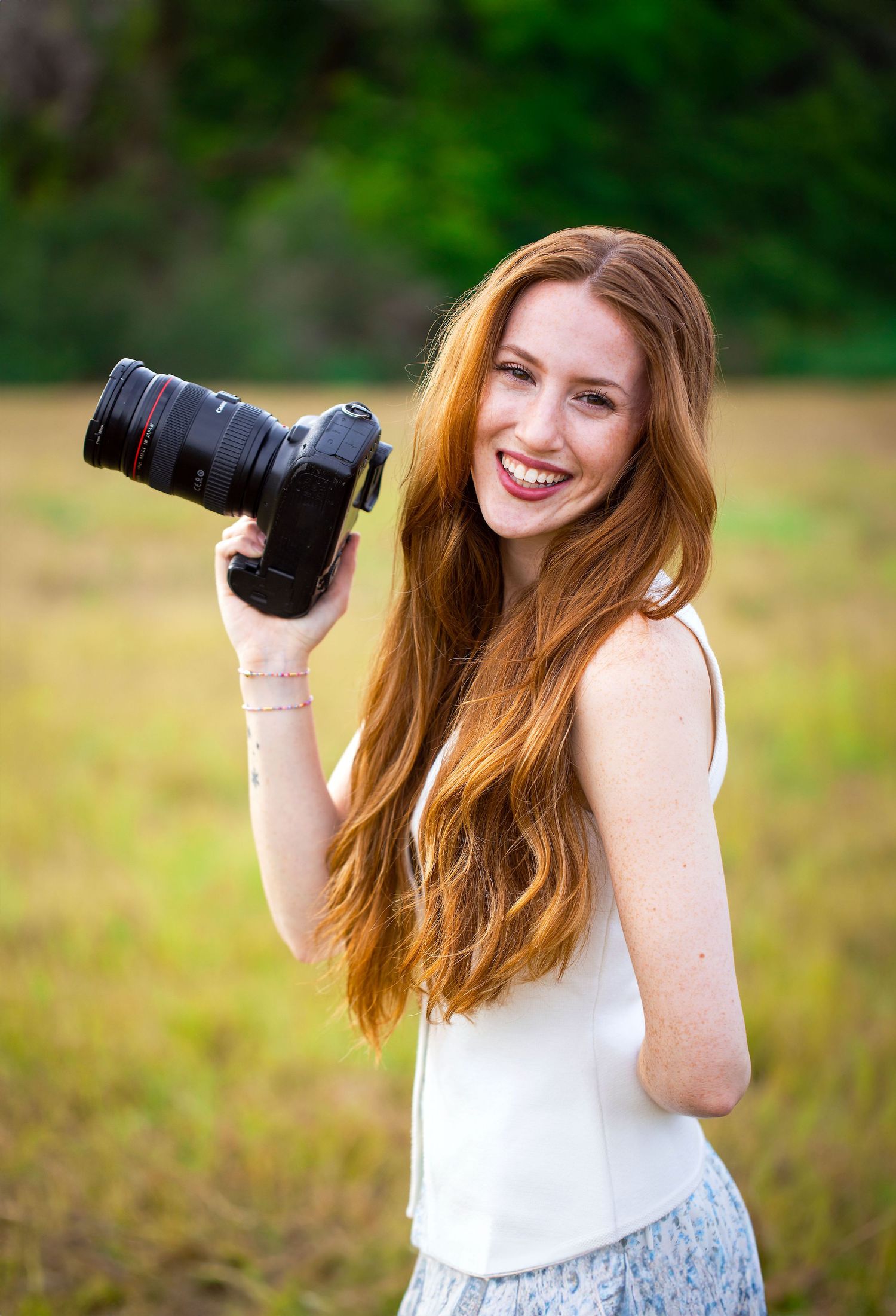 Photographer in white top holds camera while taking outdoor portraits in natural setting.