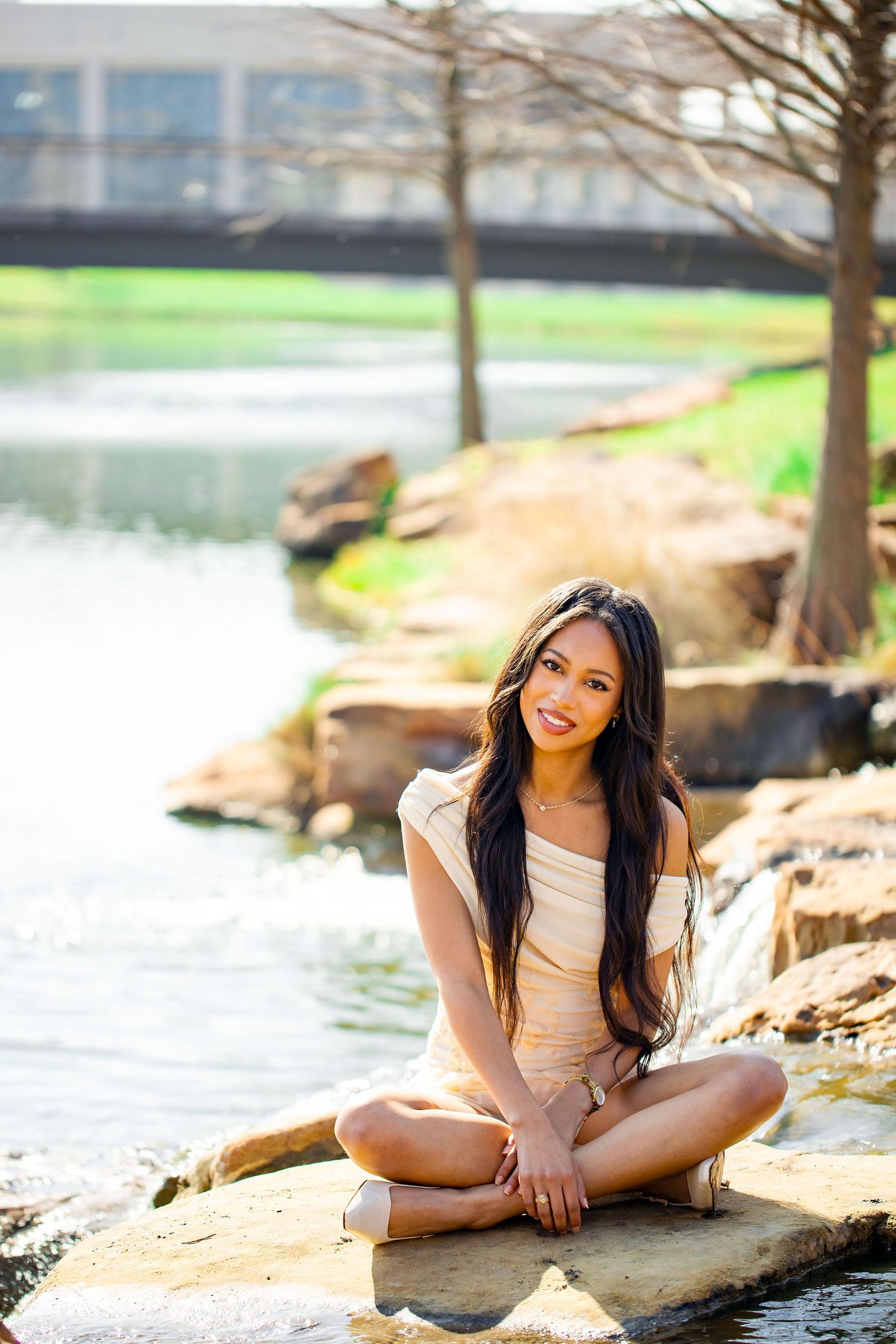 Young woman in beige outfit poses gracefully on rocky shoreline near calm water during outdoor photoshoot.