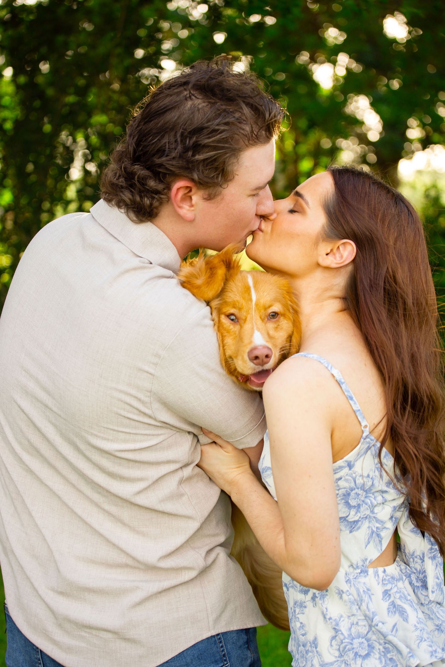 Couple kissing while holding an adorable golden dog outdoors surrounded by lush green trees.