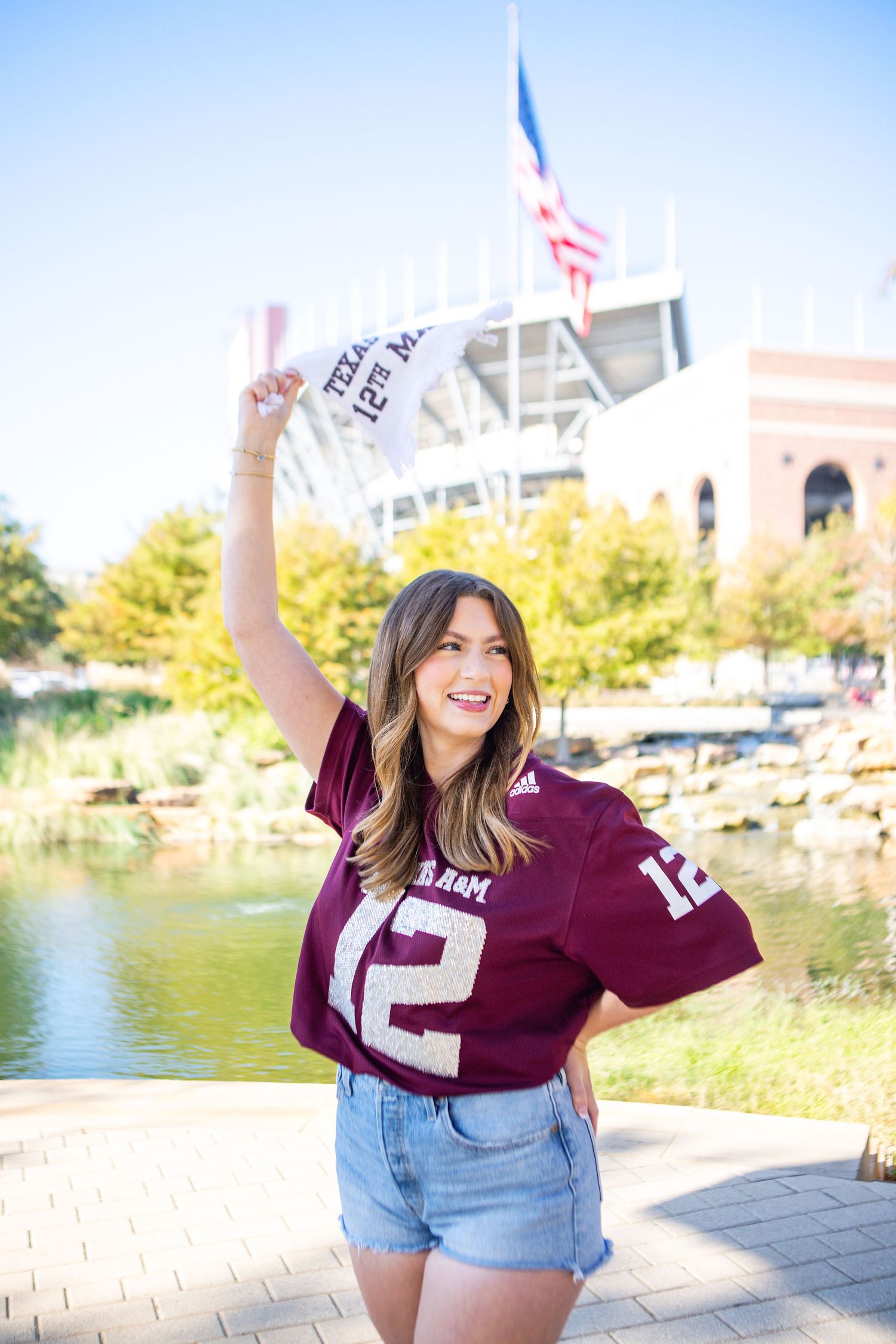 Texas A&M University graduate in maroon sports jersey and denim shorts poses at Aggie Park with stadium visible in background.