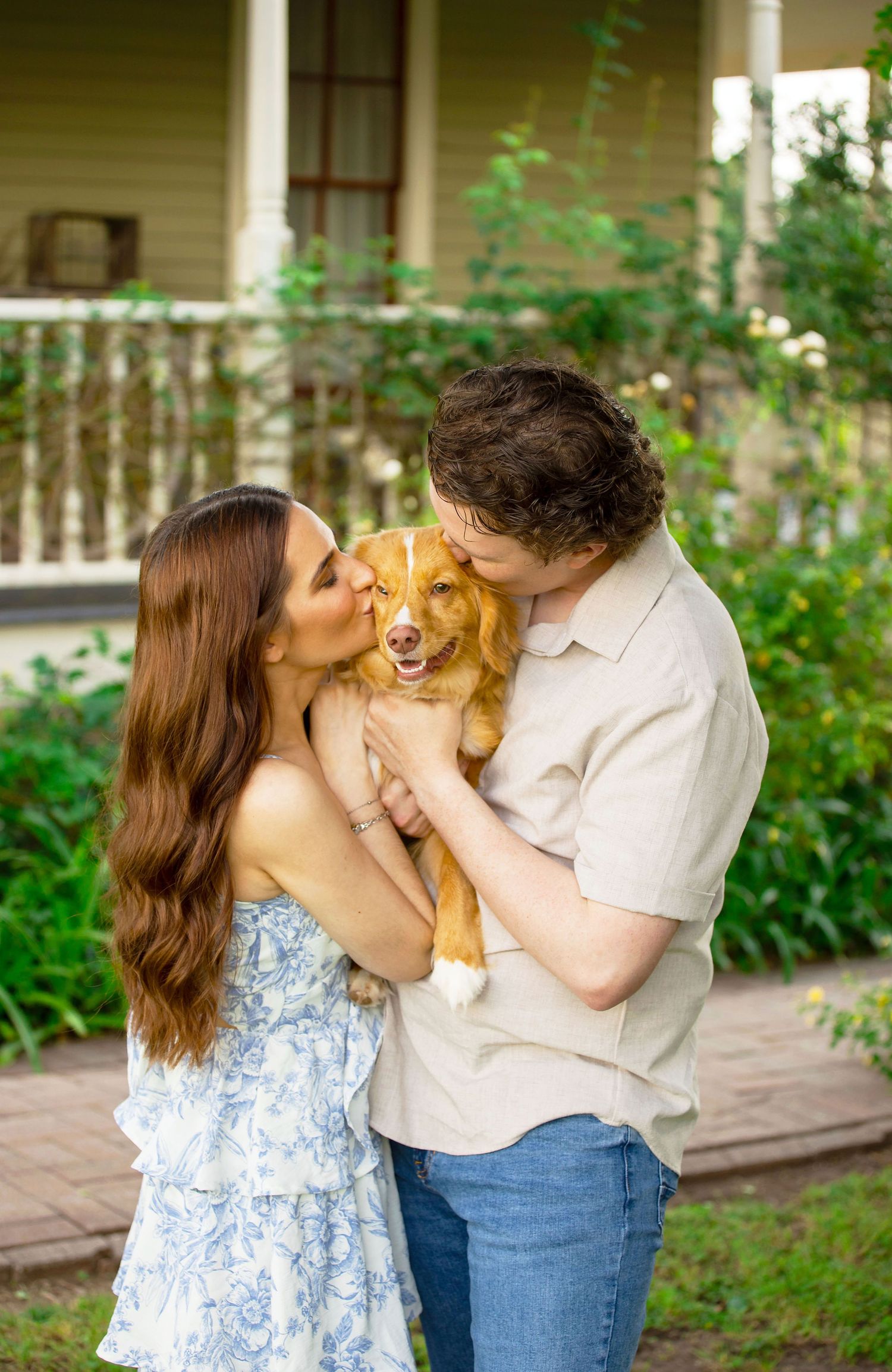 A couple kisses their golden retriever dog outdoors in front of a charming house with lush greenery.