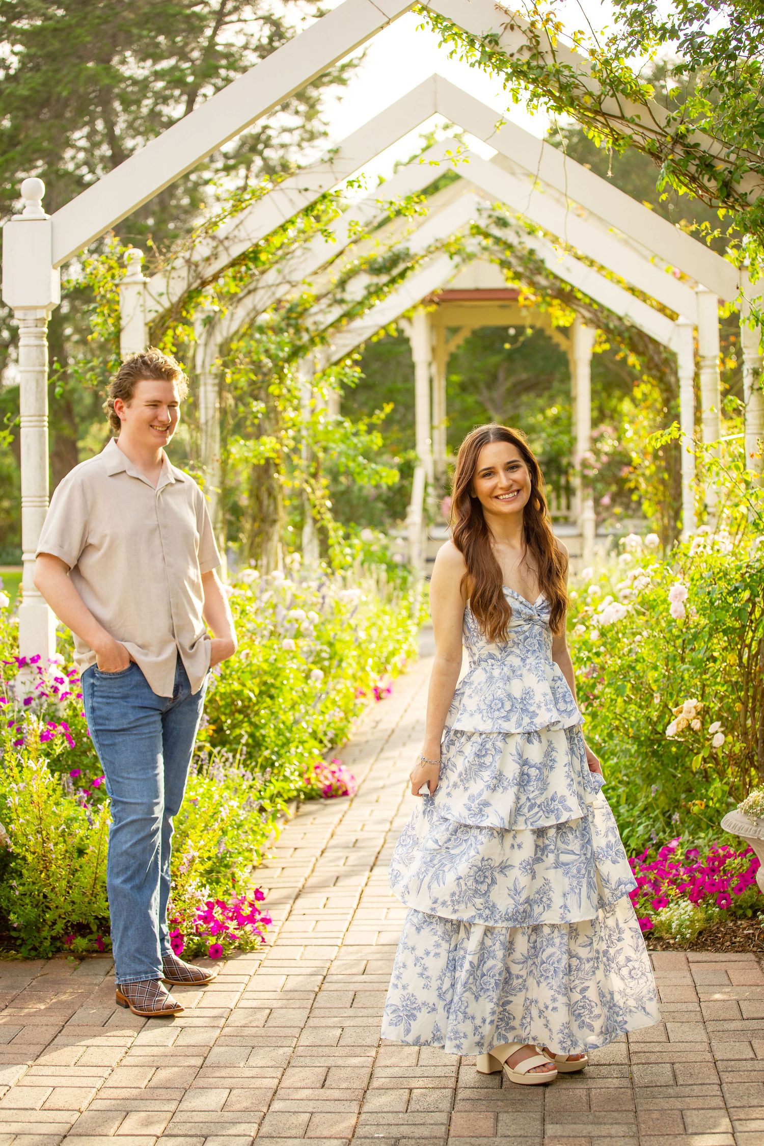 A couple walks through a white pergola garden path surrounded by colorful blooms, woman in blue floral tiered dress.