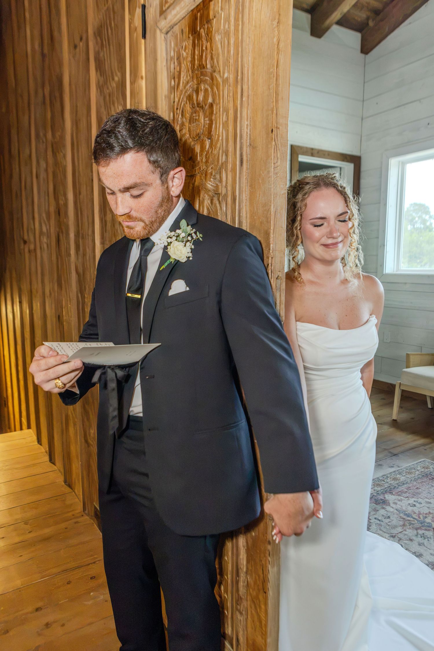 Groom reads wedding vows letter while bride smiles behind him in rustic wood-paneled venue.