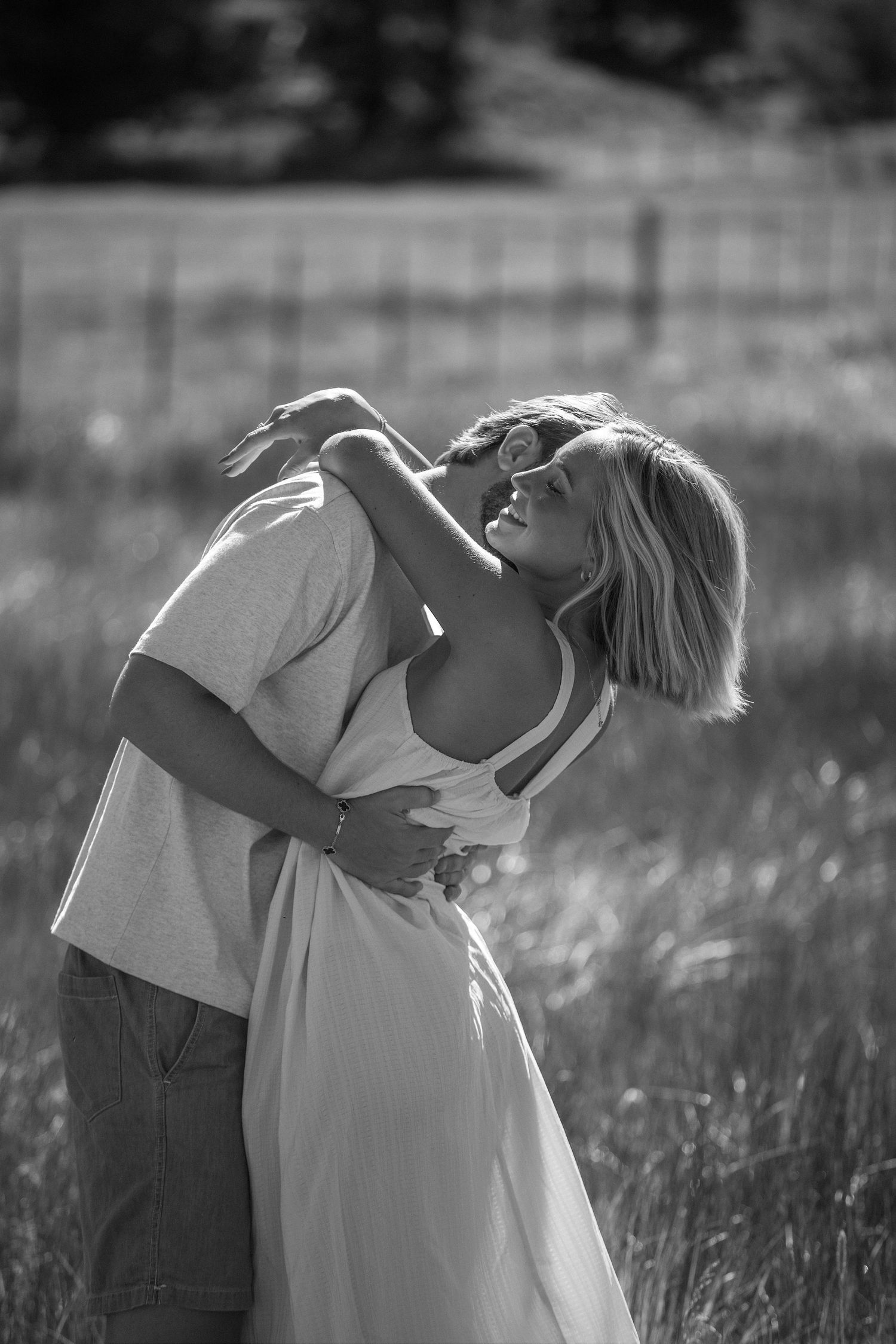 Black and white photo of a couple embracing romantically in a sunlit field, woman in white dress leaning back joyfully.
