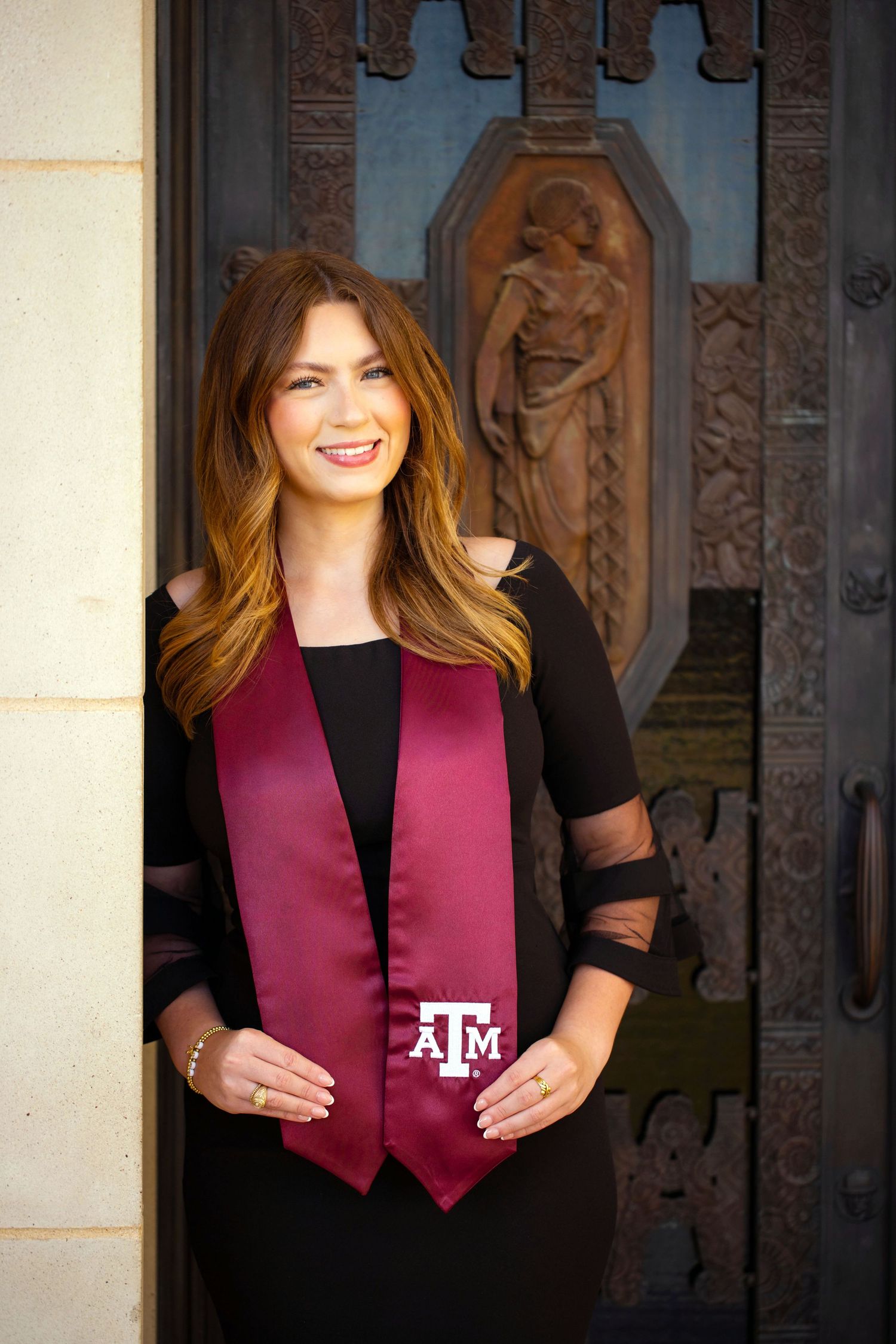 A smiling young woman wearing a maroon vest with the Texas A&M logo stands in front of an ornate stone doorway.