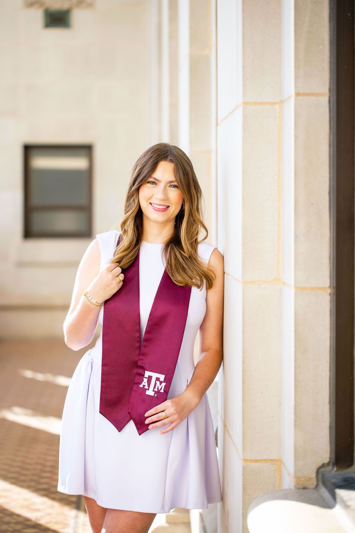 A smiling young woman wearing a maroon vest and white skirt stands in front of a building.