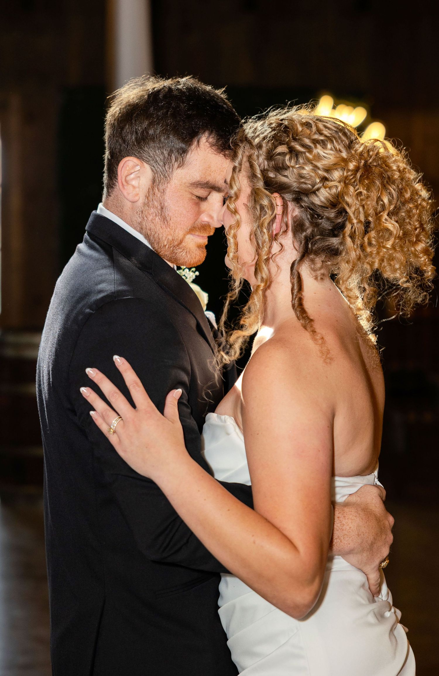 Bride and groom share an intimate first dance moment, foreheads touching, dressed in wedding attire at a reception venue.