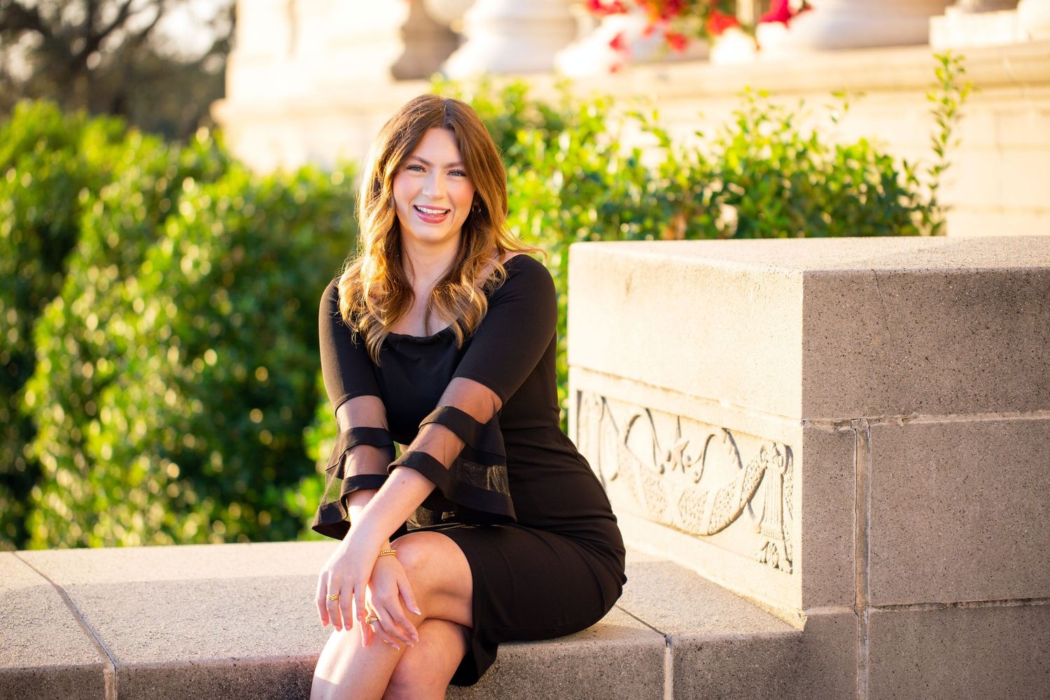 A young woman with long brown hair is sitting on a concrete wall, smiling and wearing a black outfit.