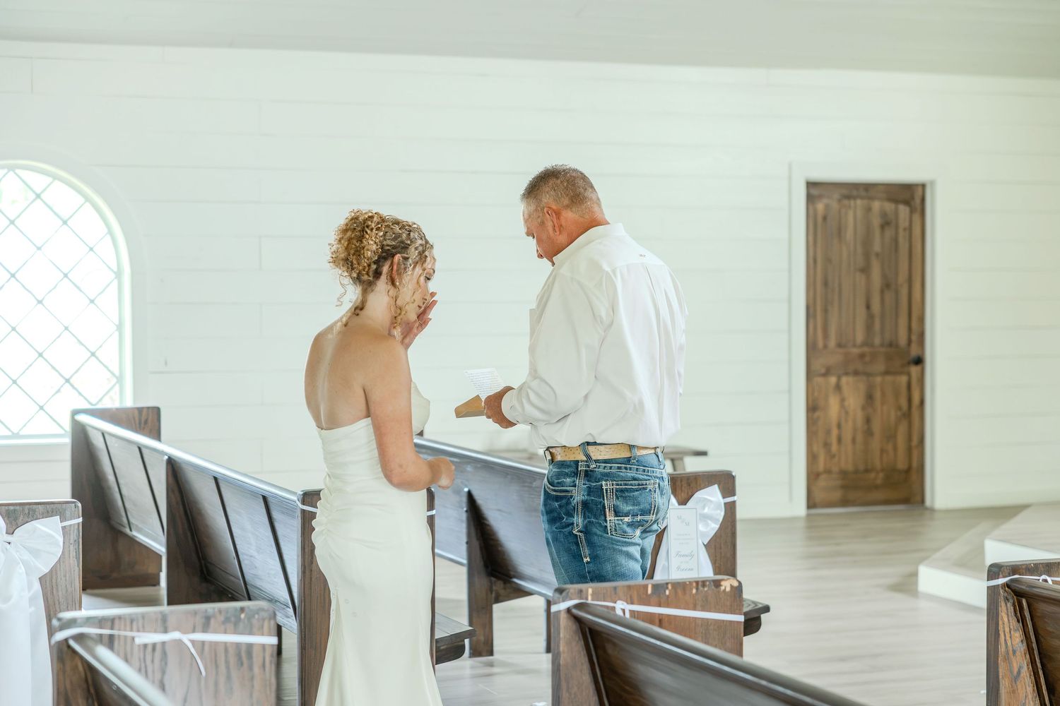 Bride and groom stand together at the altar inside a rustic white chapel with wooden pews during their wedding ceremony.