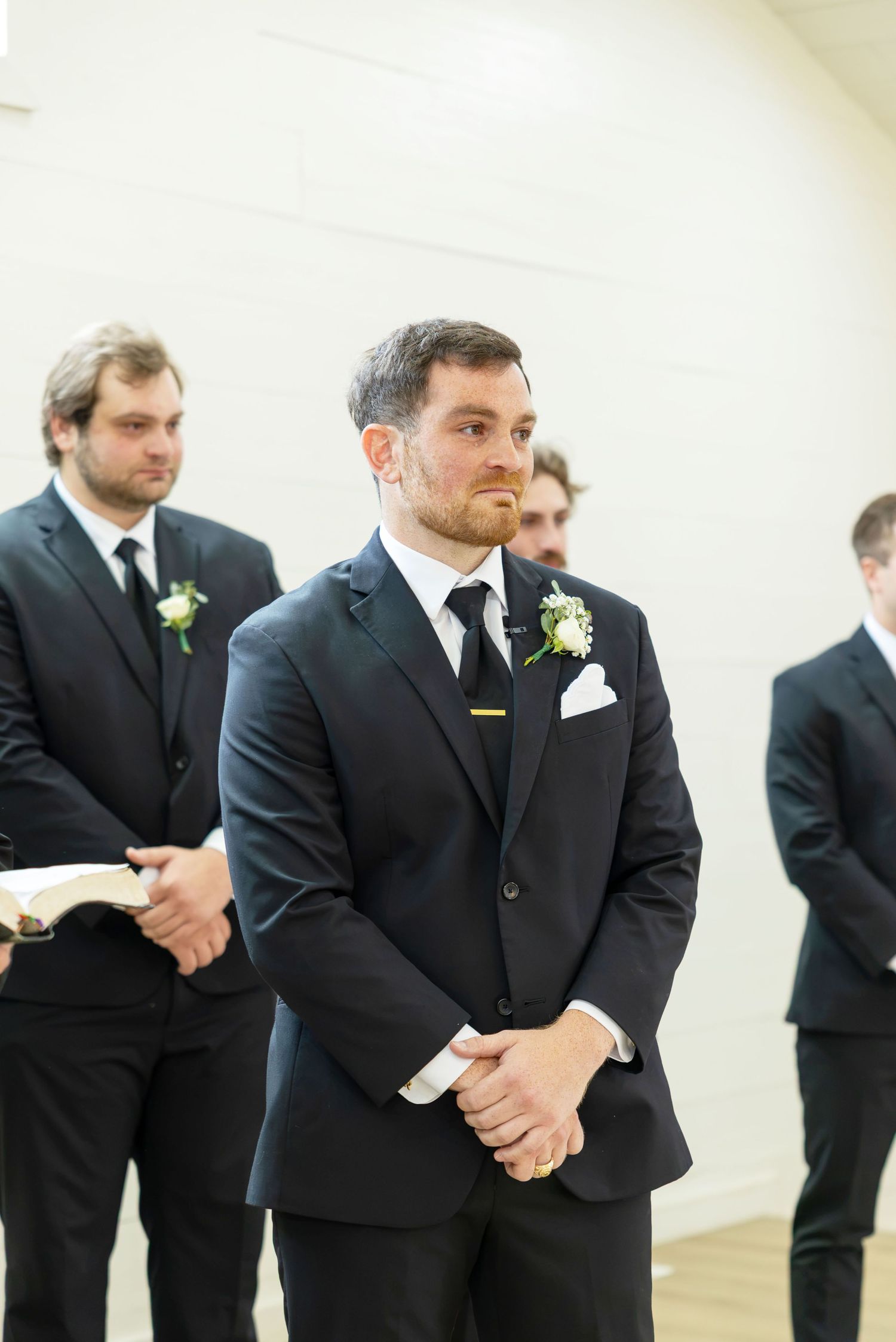 Groom in dark suit with white boutonniere stands at altar with groomsmen during wedding ceremony.