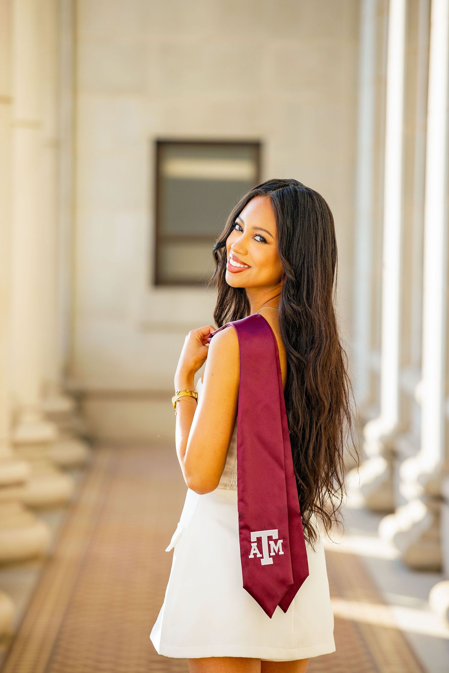Graduate in white dress poses with maroon Texas A&M tie in elegant columned hallway.