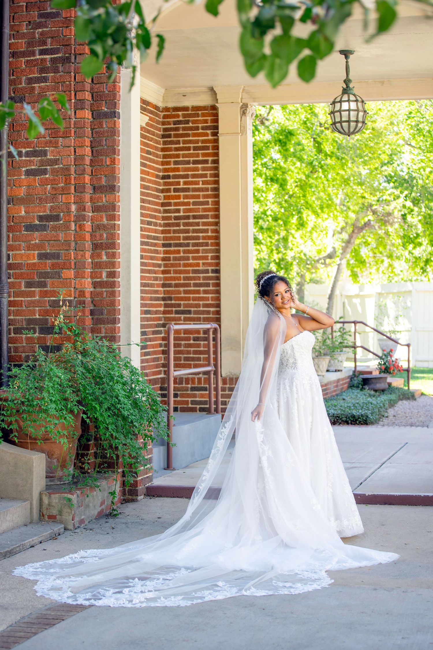 Bride in Coreena's Bridal lace wedding gown with cathedral veil poses under brick columned porch.