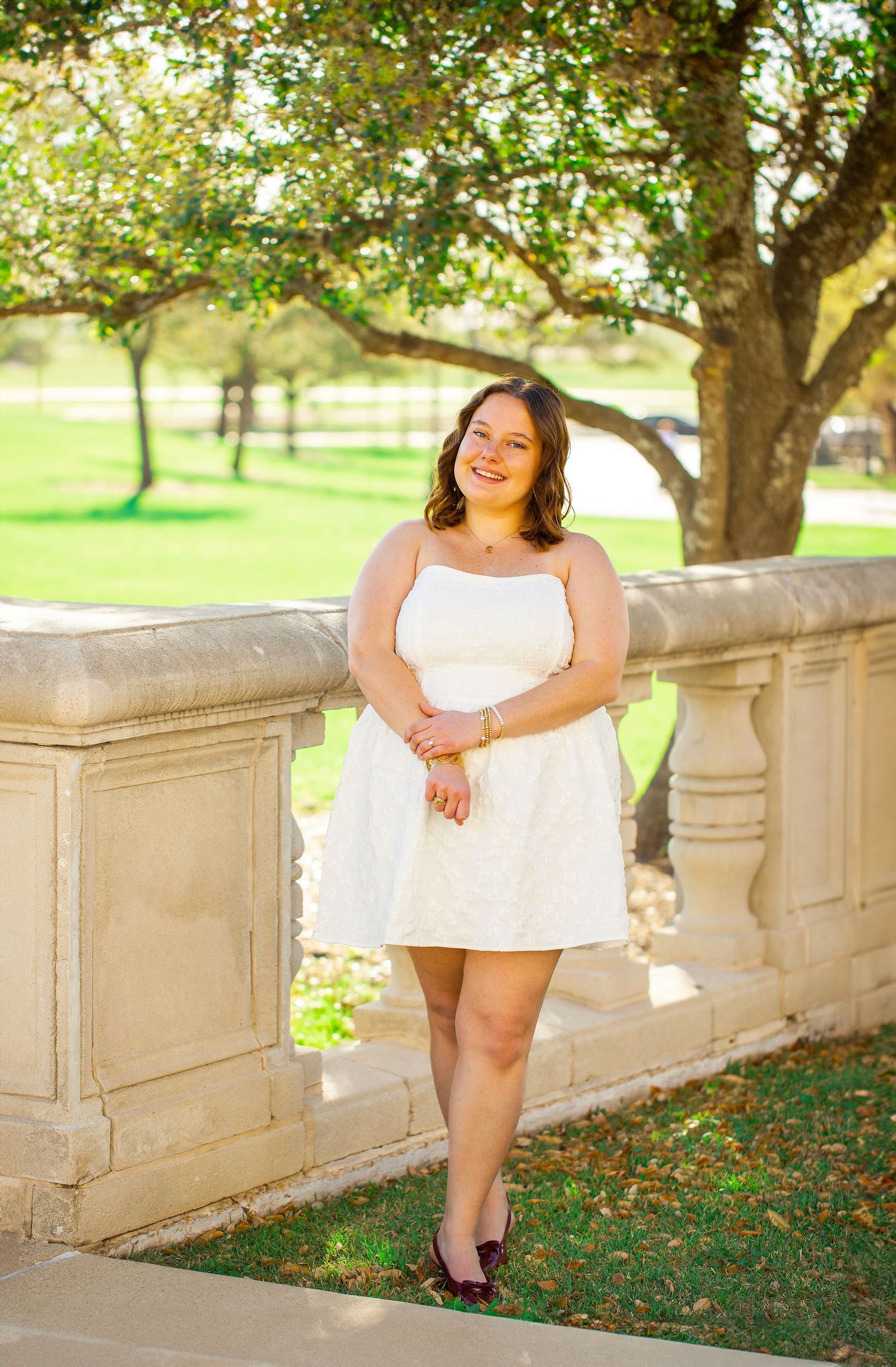 Young woman in white sundress smiling outdoors near stone balustrade with green trees in background.