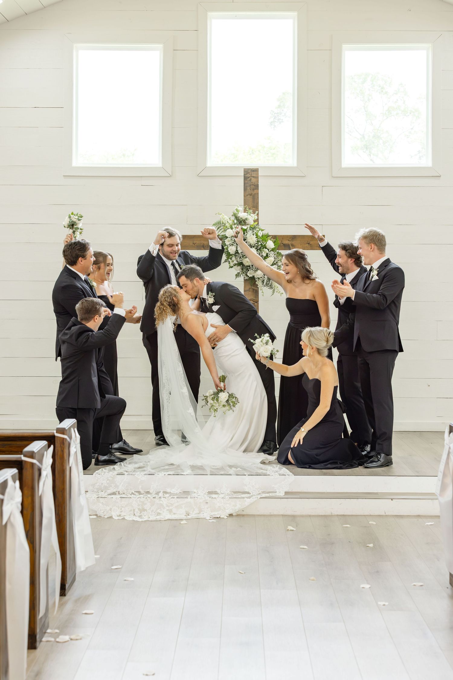 Bride and groom share first kiss surrounded by wedding party in black attire inside a white chapel with wooden cross.