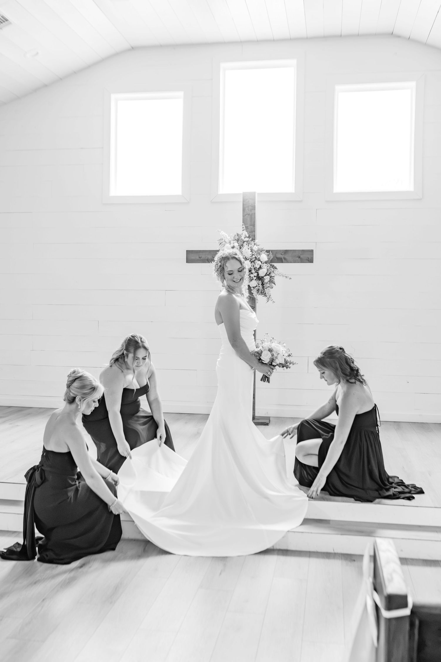 Bride in white gown with bridesmaids arranging her train in a chapel with a wooden cross and floral arrangement.