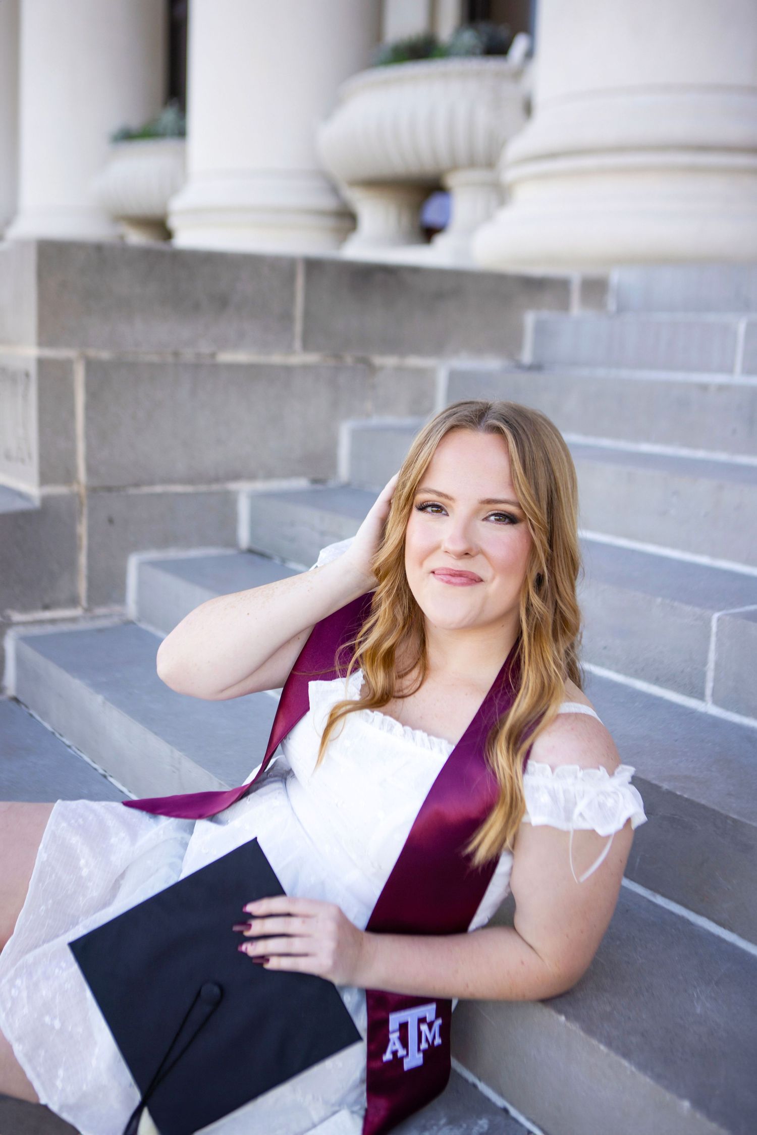 Graduate poses on stone steps wearing a white dress and maroon graduation stole from Texas A&M University.