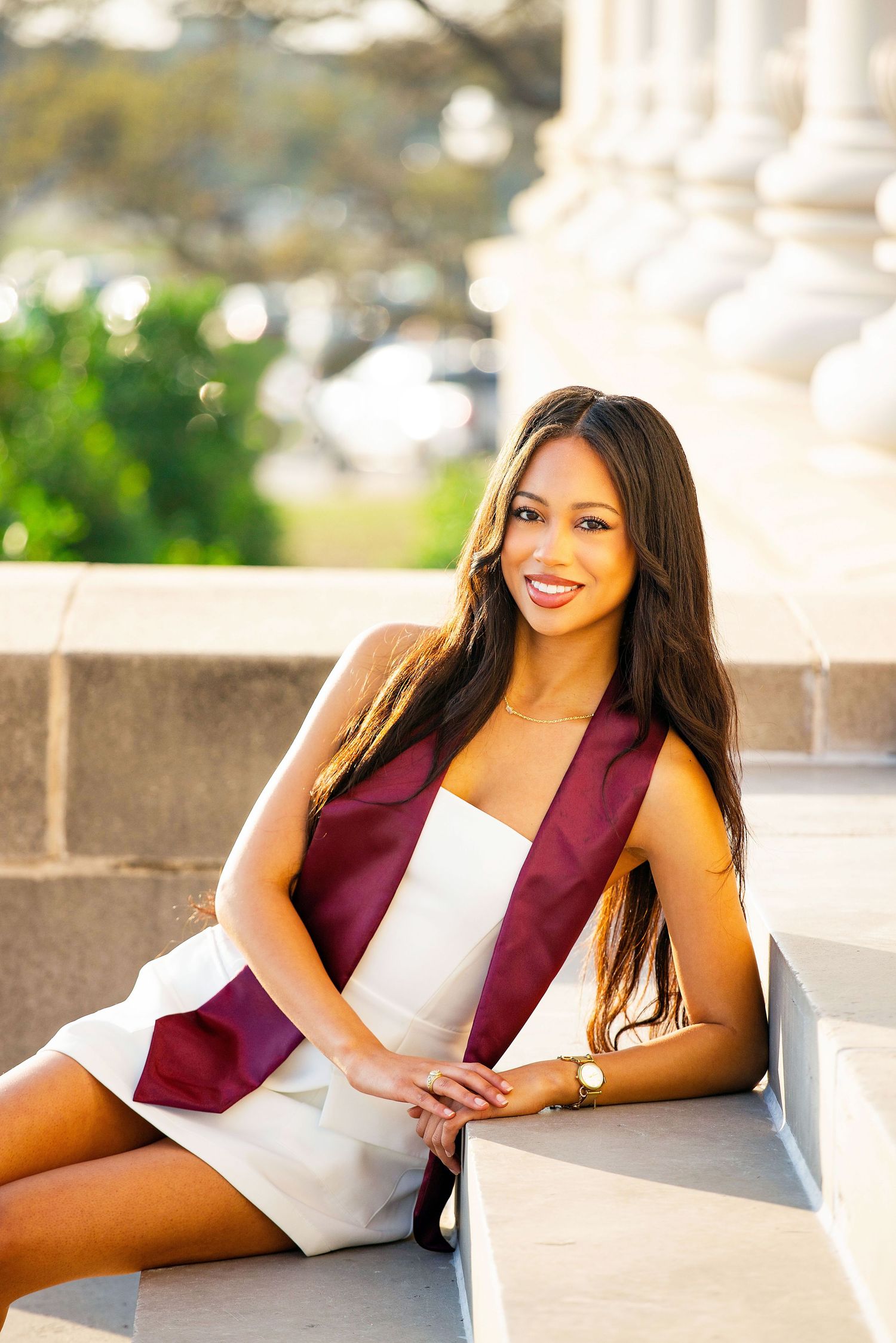 Graduate in white dress and maroon stole sits on stone steps with white columns and greenery behind.