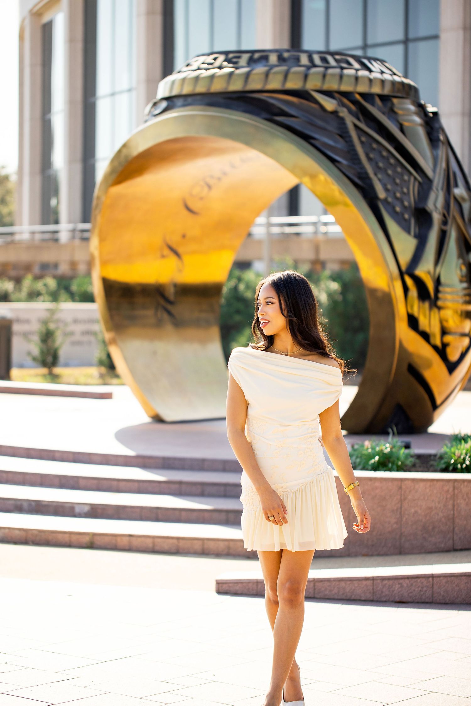 Graduate in white dress twirls joyfully in front of large golden sculpture during celebration photoshoot.