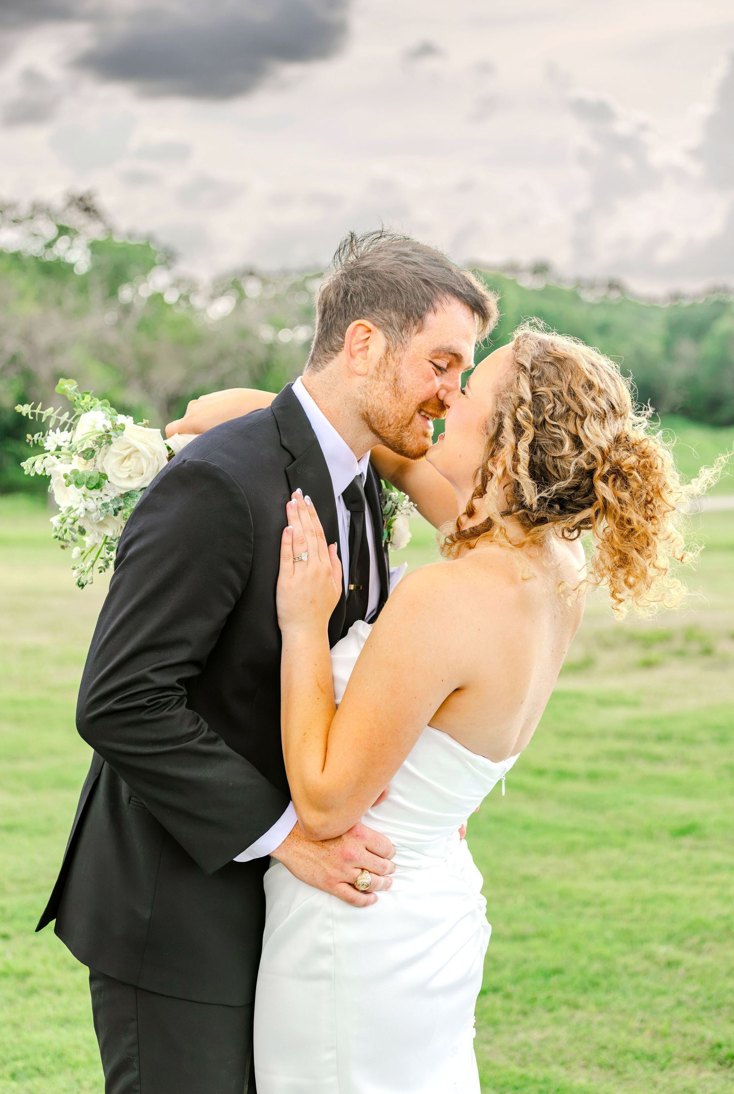 Bride and groom share a romantic first kiss outdoors on their wedding day, with lush green grass and dramatic clouds behind them.