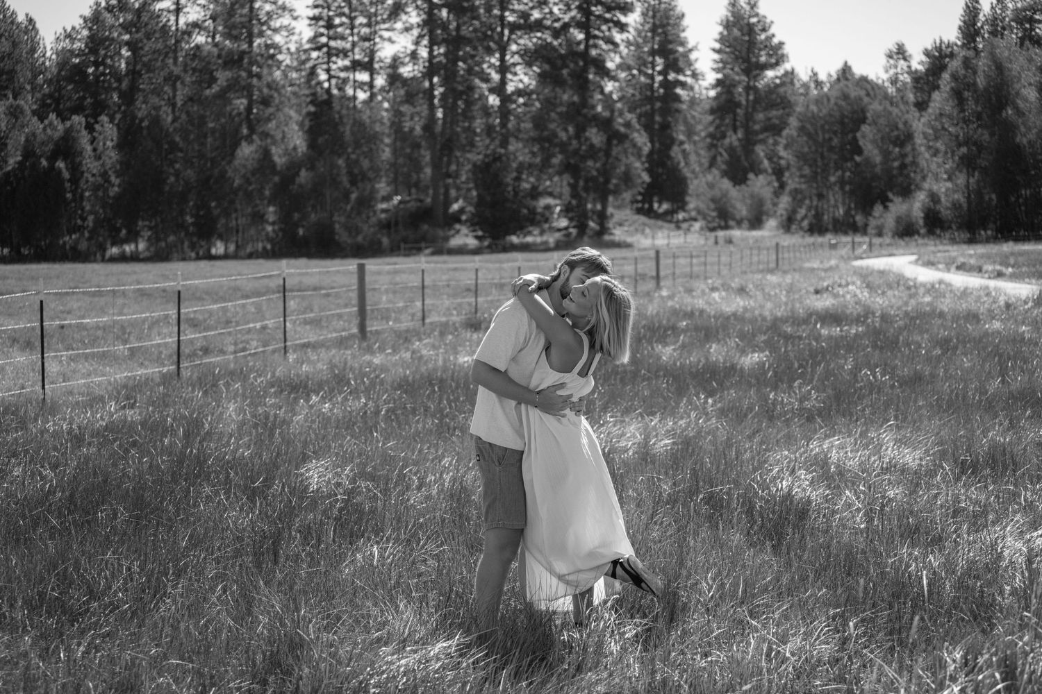 Black and white photo of a couple sharing a romantic kiss in a grassy meadow with pine trees in the background.