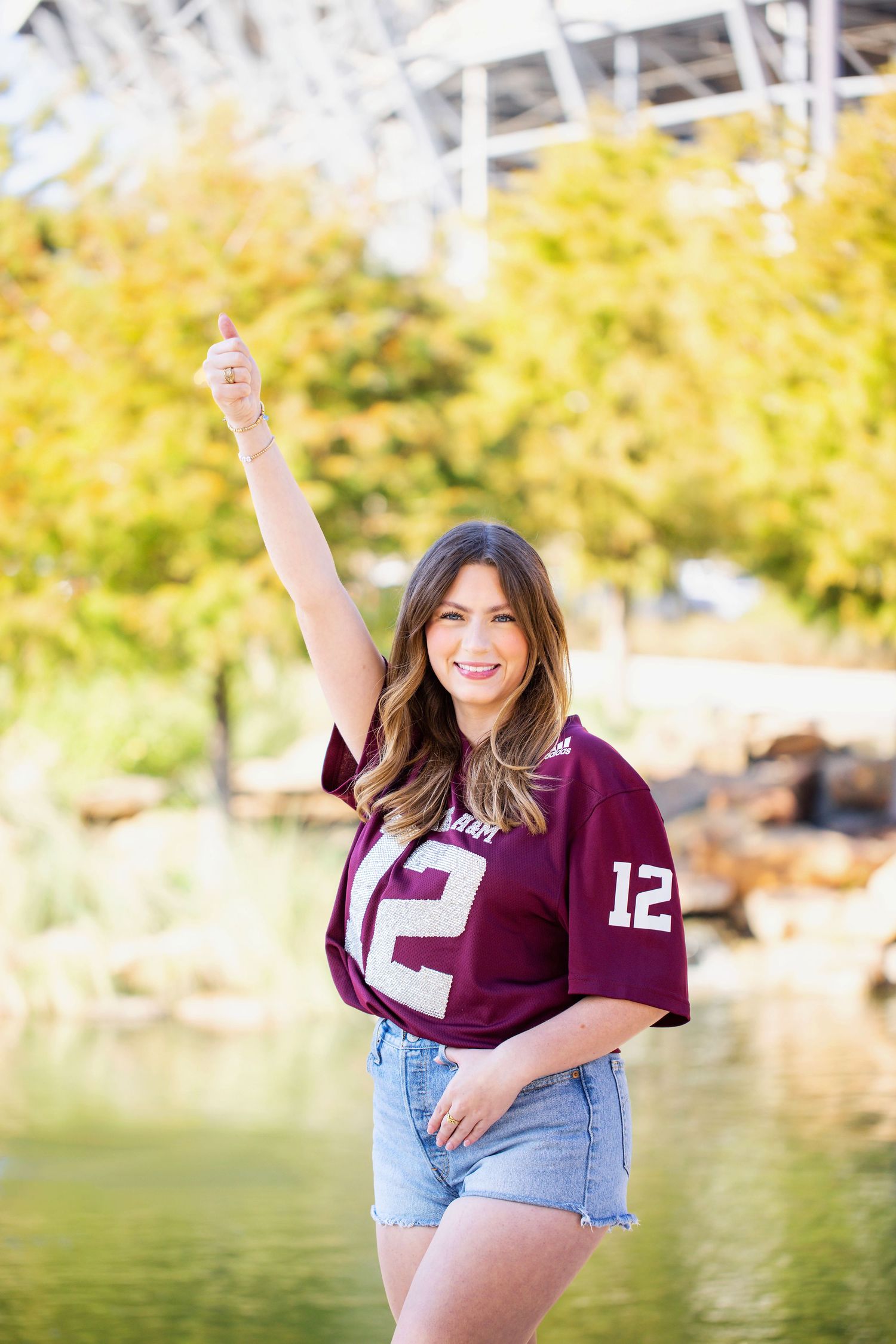 A smiling young woman in a burgundy jersey with the number 12 on it stands outdoors with her arm raised.
