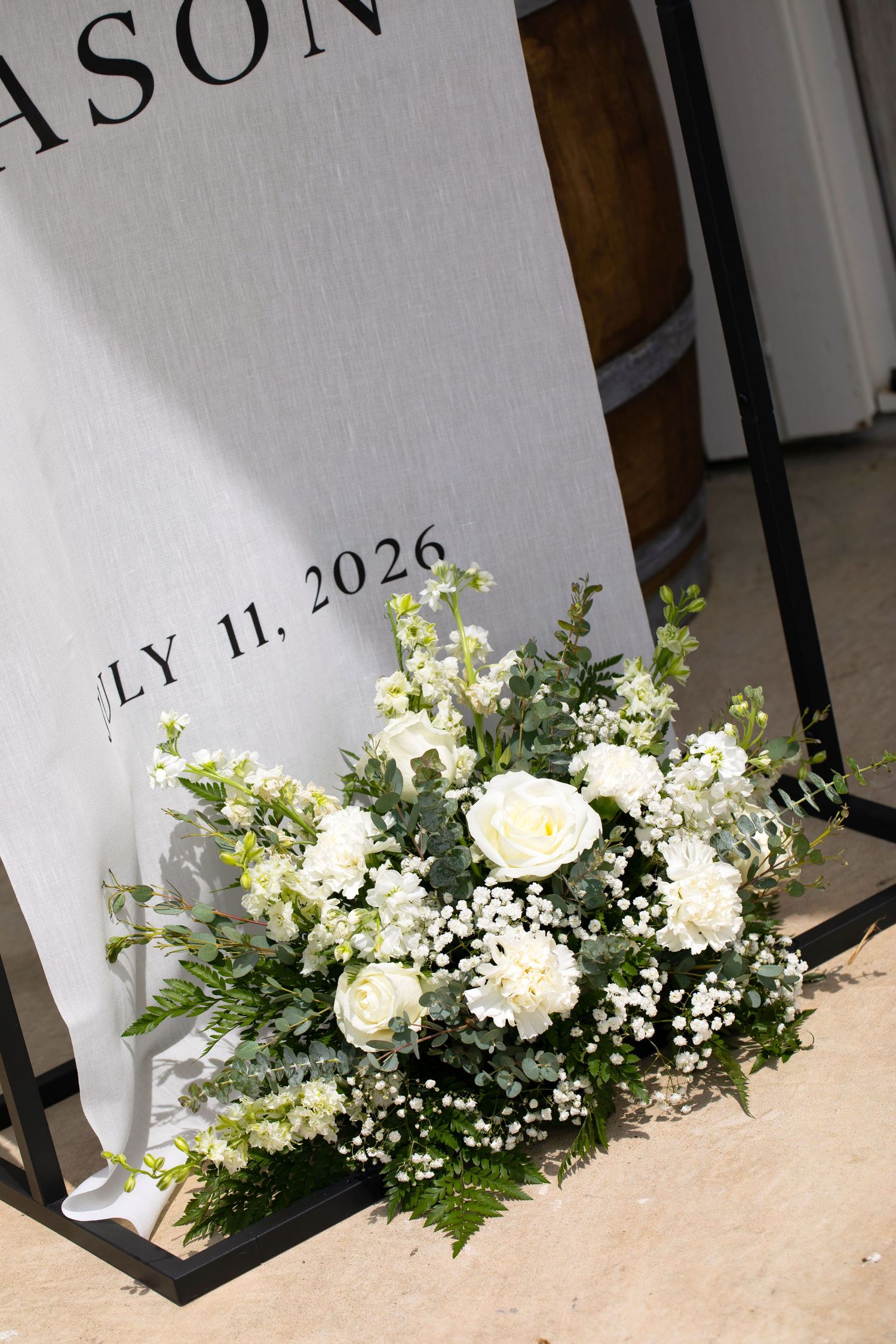 White floral arrangement with roses and greenery beside a wedding welcome sign dated July 11, 2026.