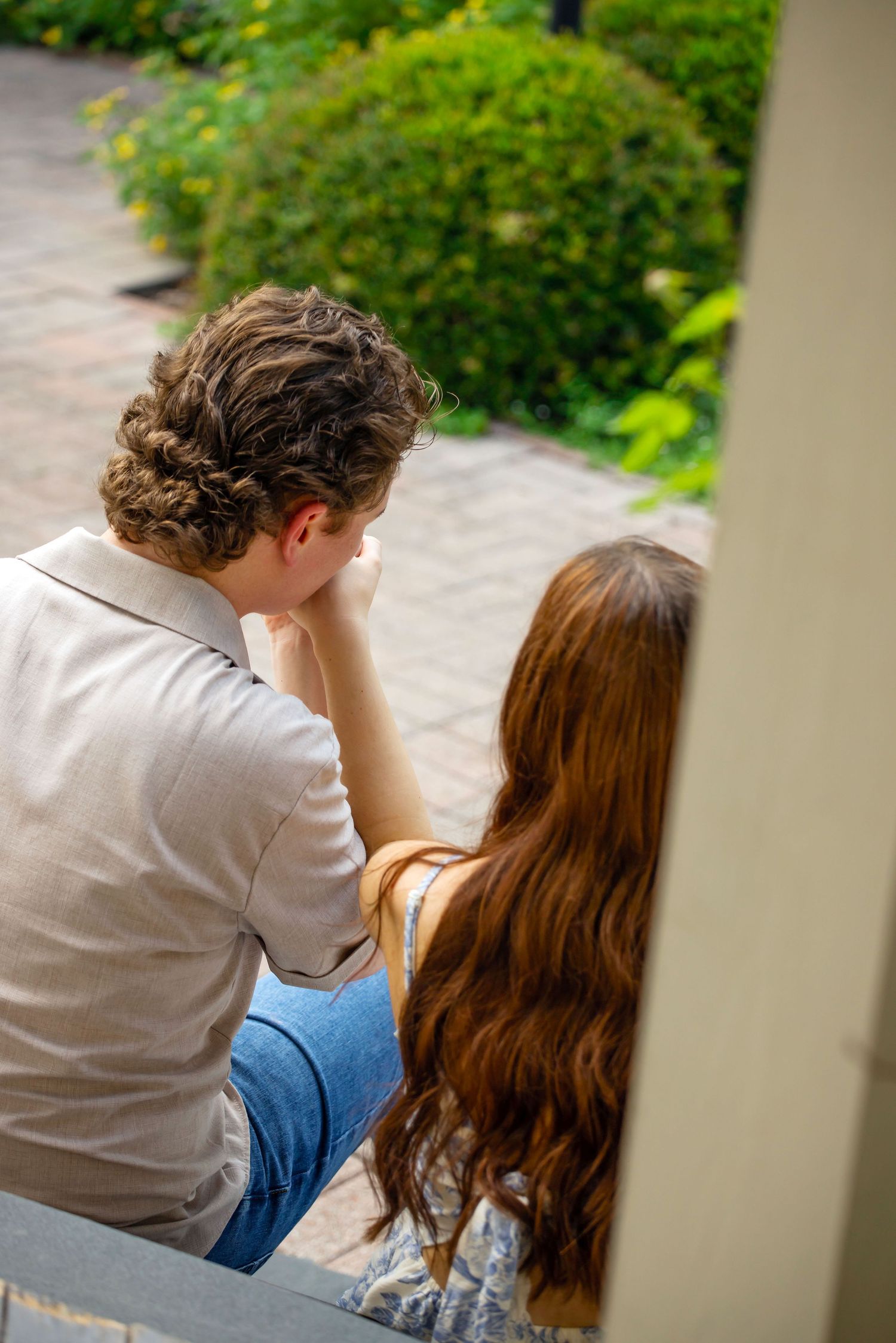 A young couple sits together on steps, viewed from behind, gazing at a lush green garden.