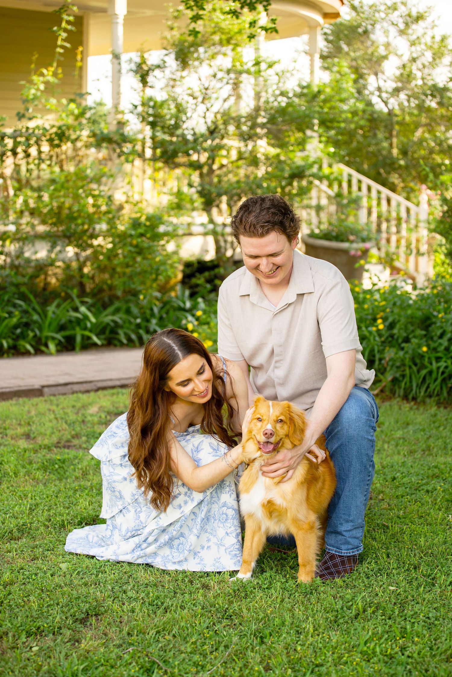 A smiling couple petting a happy golden dog on green grass in front of a charming house.