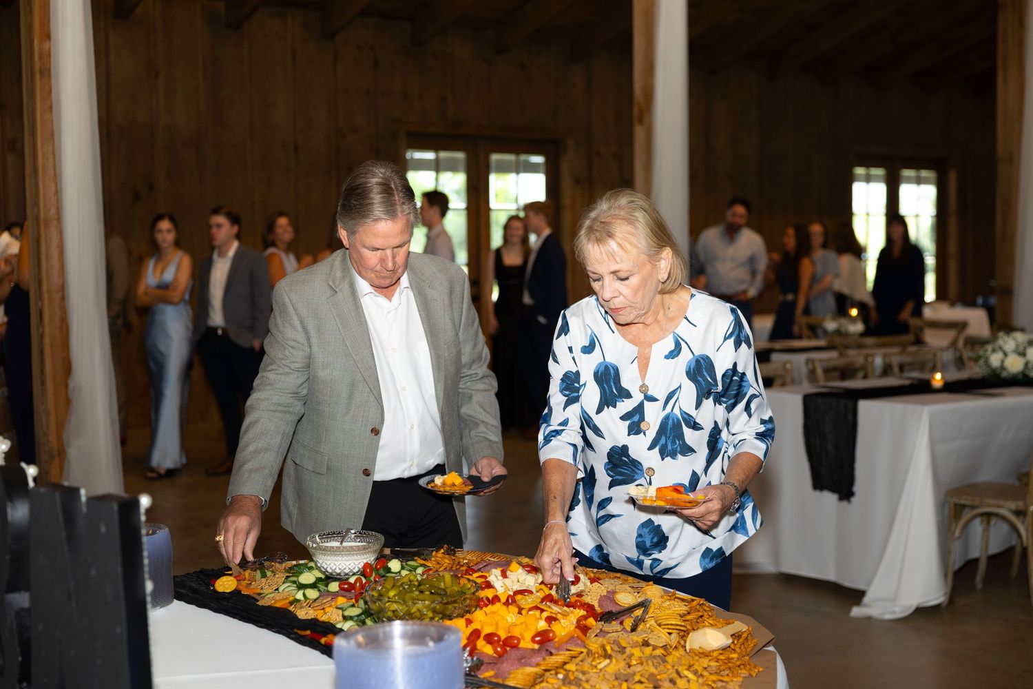 Guests serving themselves at an elegant buffet table filled with colorful dishes at an indoor barn wedding reception.