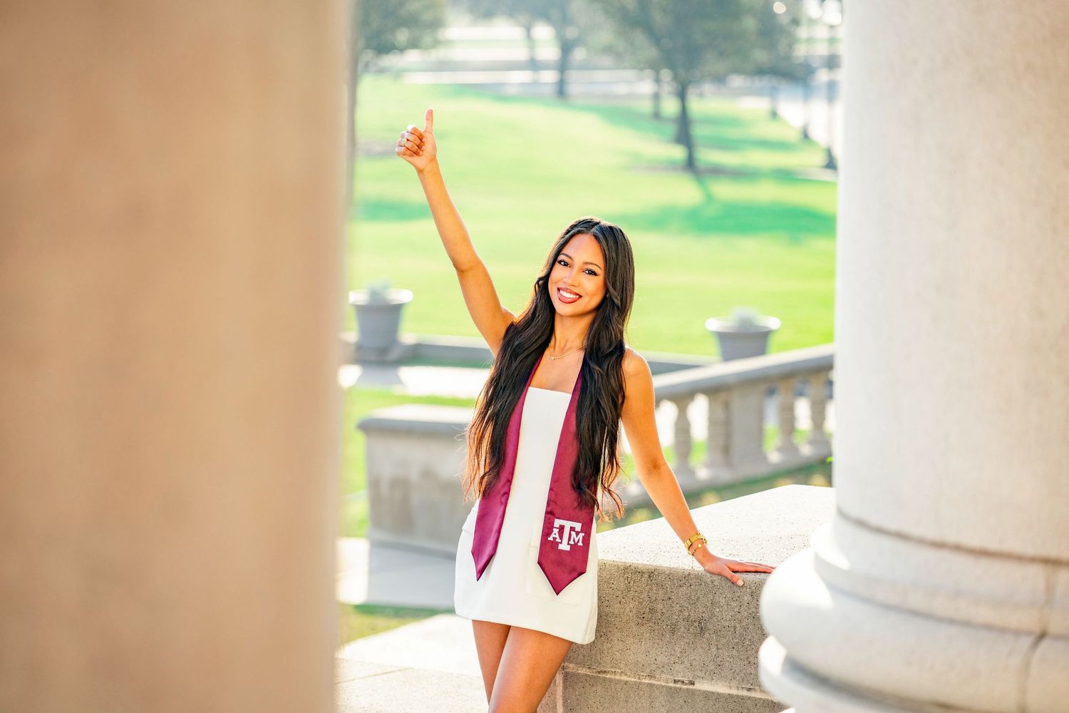Woman in a white dress poses gracefully against stone columns in a sunlit outdoor setting.