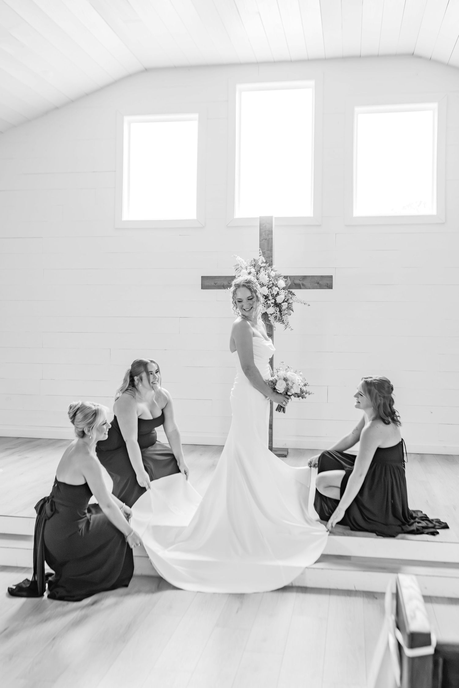 Bride in white gown stands before wooden cross as bridesmaids in dark dresses arrange her train in a chapel.