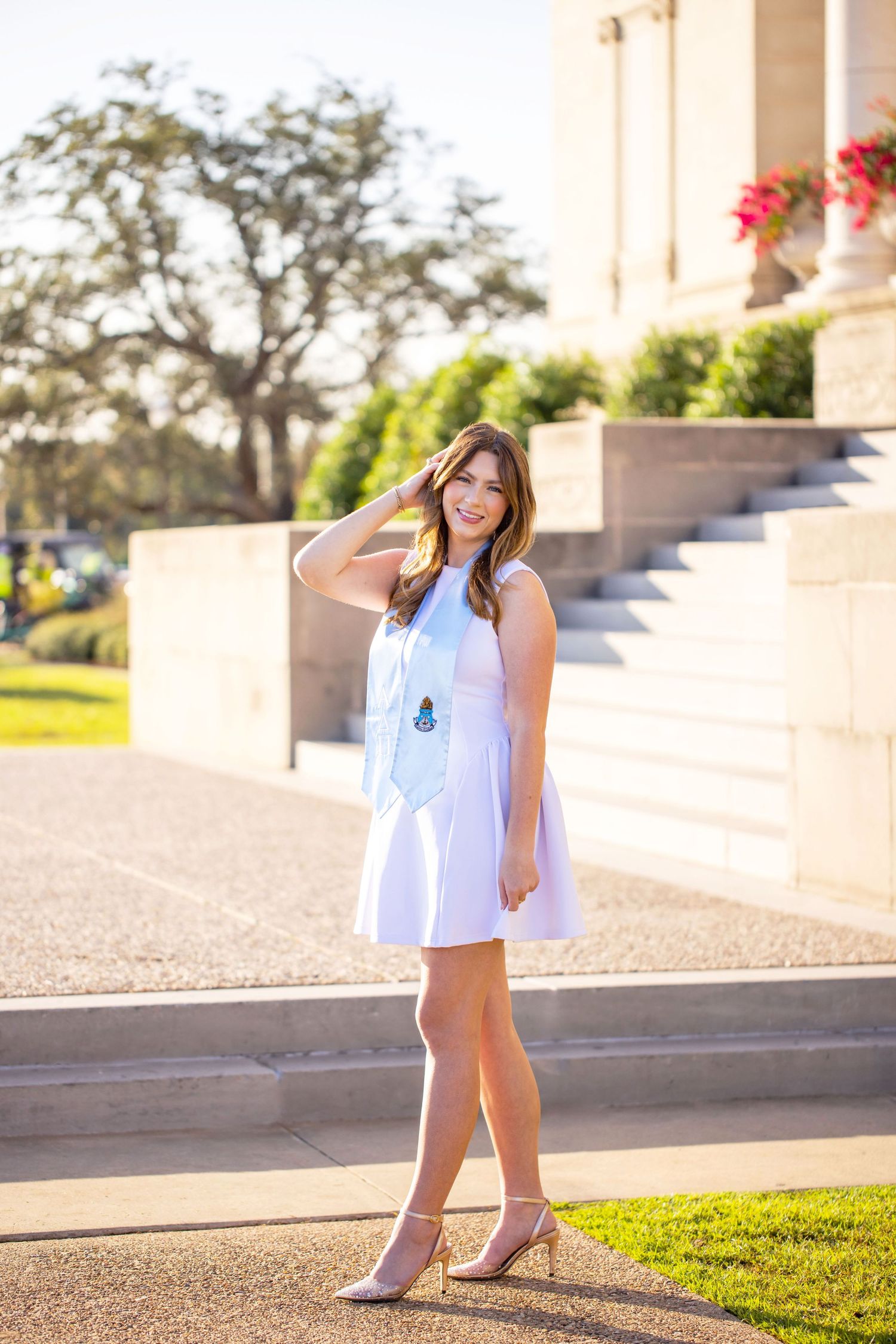 A smiling woman in a white dress stands on a grassy path near a building with flowers.