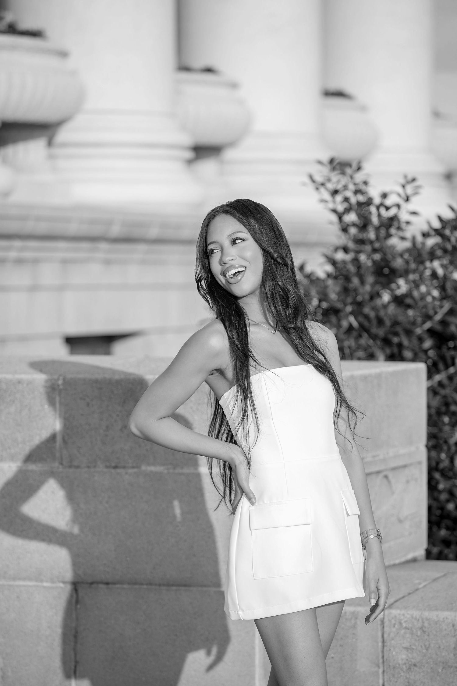 A person in a white summer dress poses against classical stone columns in a black and white photograph.