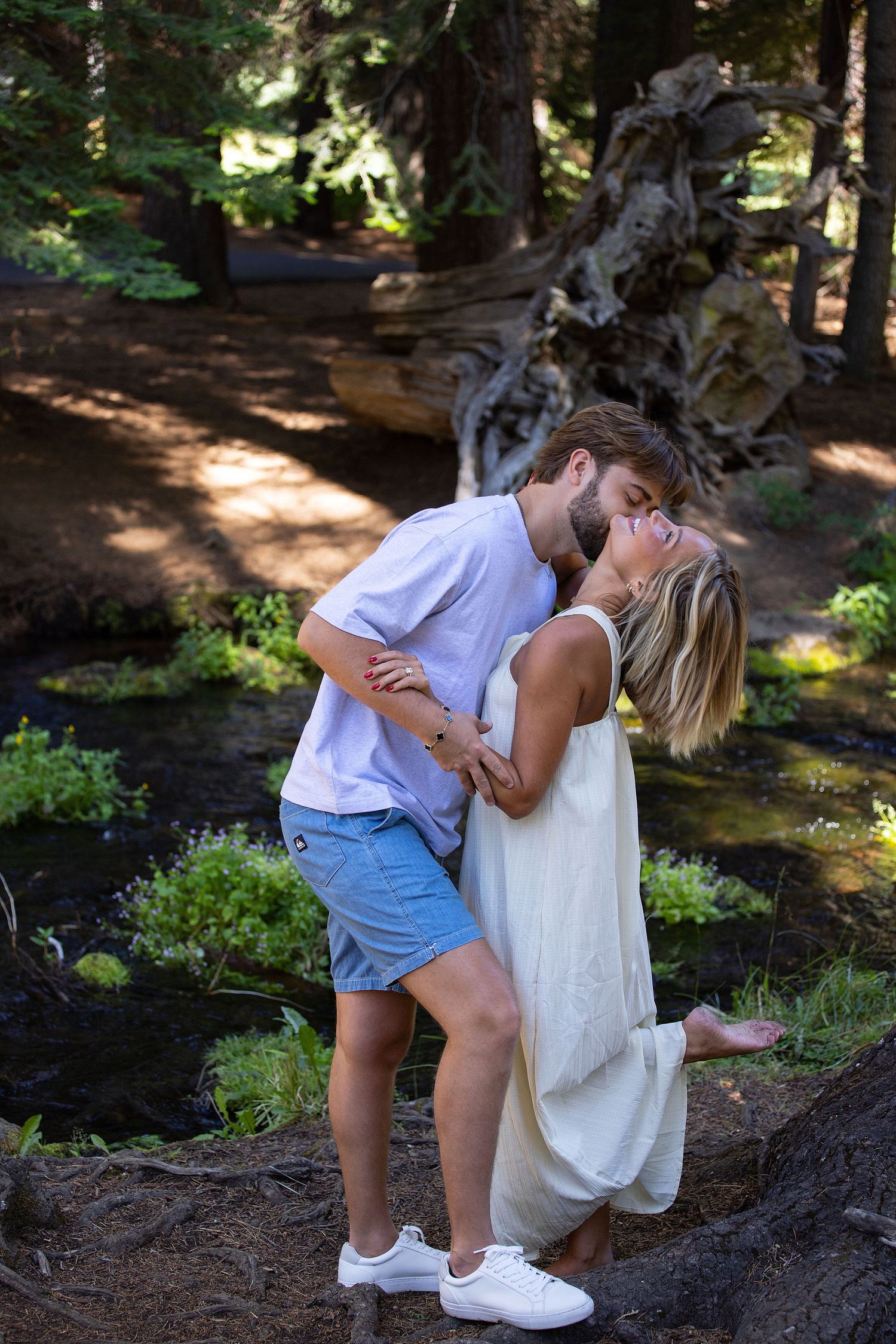 Couple sharing a romantic kiss in a lush forest setting, man in blue shorts and woman in white dress.