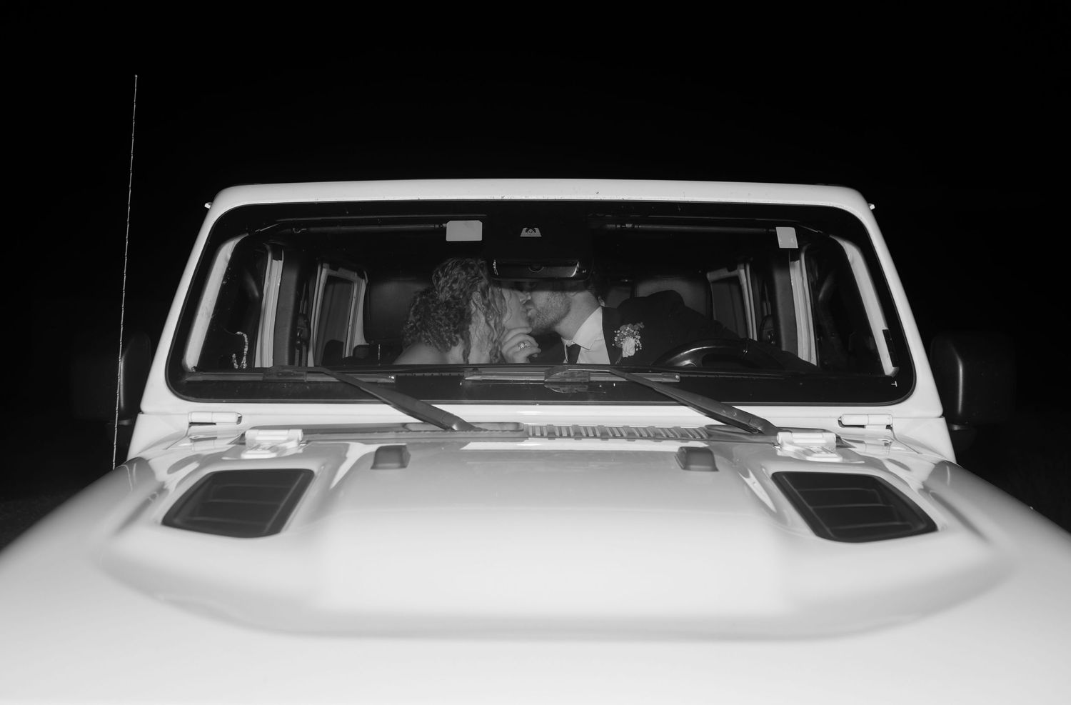Black and white photo of people inside a white Jeep Wrangler viewed through the windshield at night.