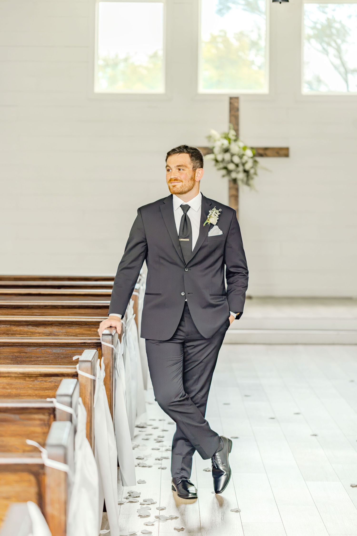 Groom in dark suit with boutonniere stands confidently in white chapel aisle beside wooden pews before wedding ceremony.