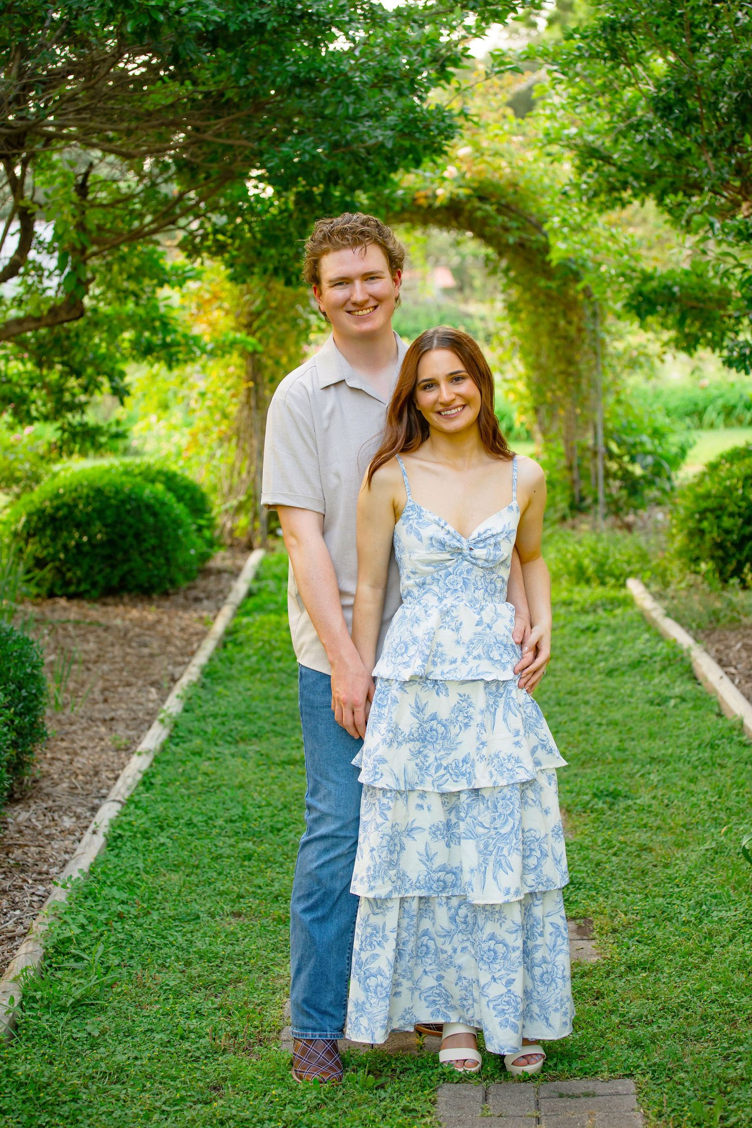 Young couple smiling together in a lush garden pathway, woman in blue floral tiered dress, man in casual shirt and jeans.