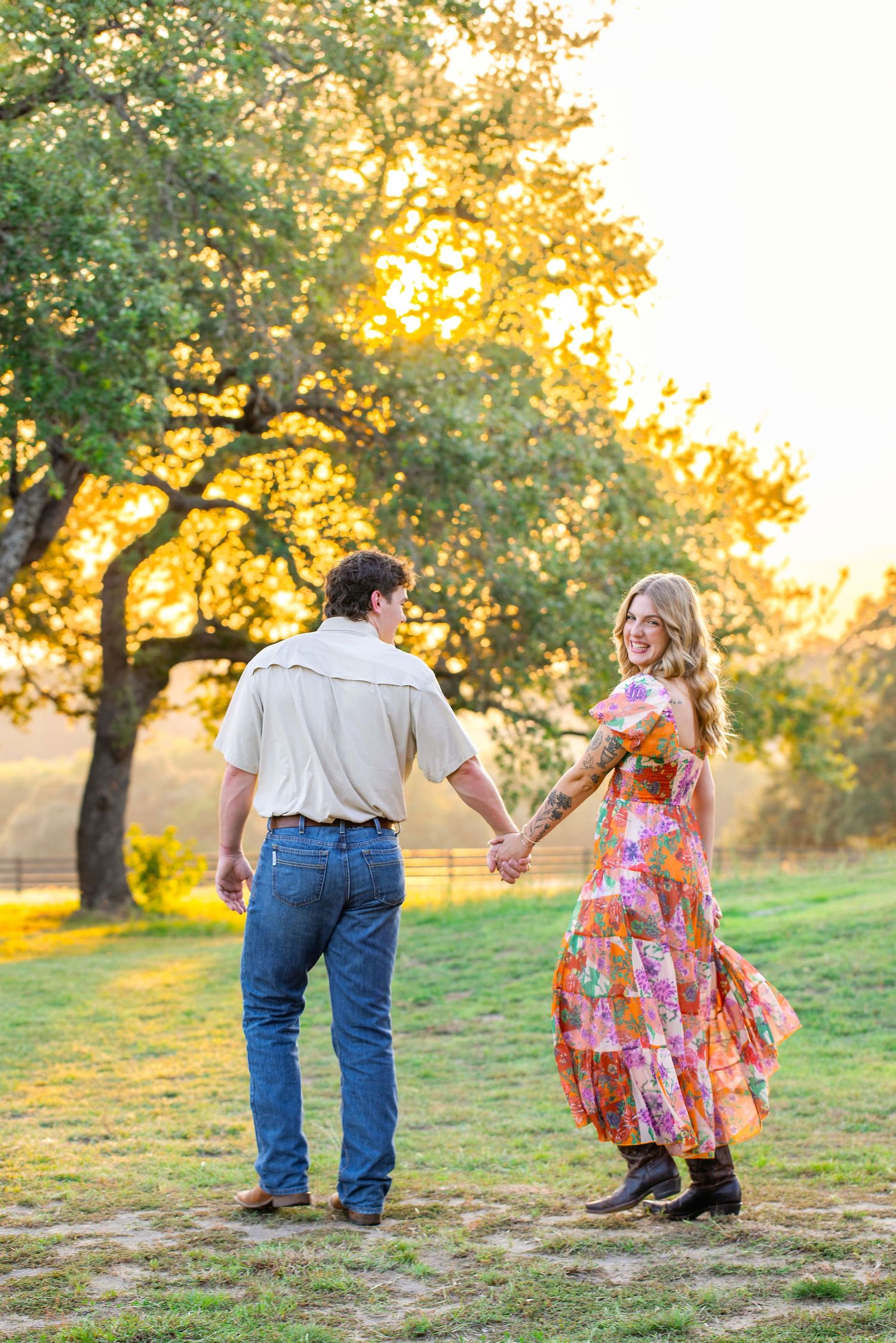 Couple holding hands while walking through a sunlit meadow at golden hour with trees in the background.