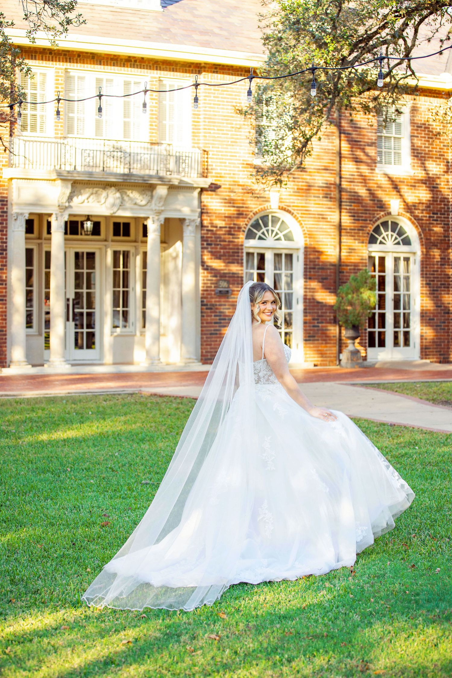 A bride in a Coreena's Bridal flowing white wedding gown poses in front of a grand brick mansion with arched windows.