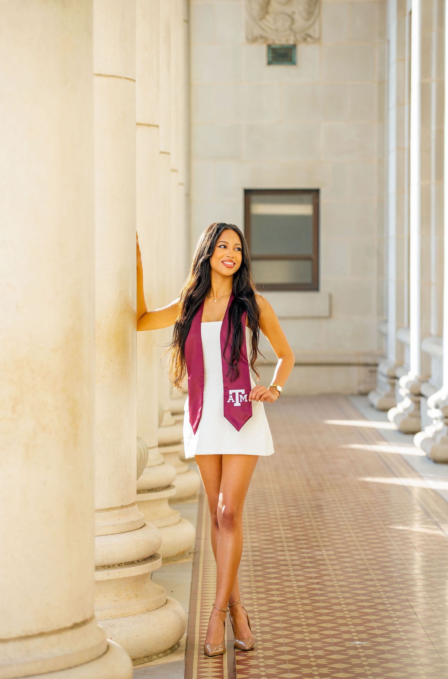 Graduate in white dress and maroon Texas A&M stole poses elegantly between classical white columns.
