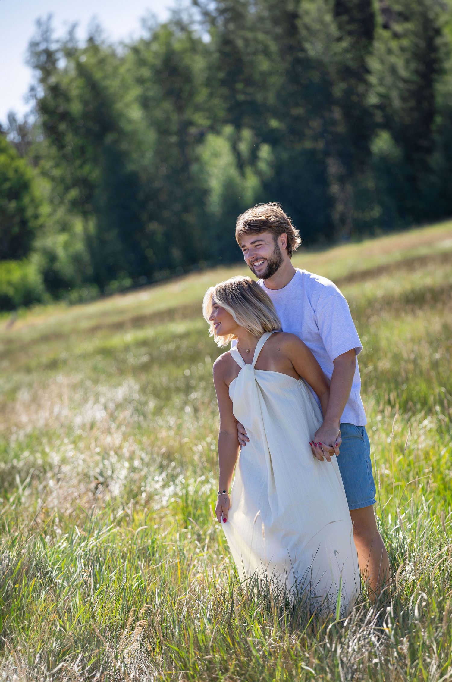 A couple embraces in a sunlit meadow, the woman in a white dress and man in a white tee, with lush green trees behind.