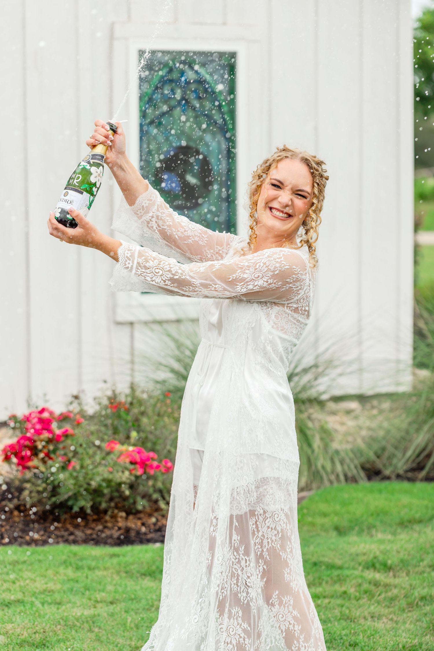 Smiling bride in white lace gown popping champagne bottle outdoors near white barn with colorful flowers.