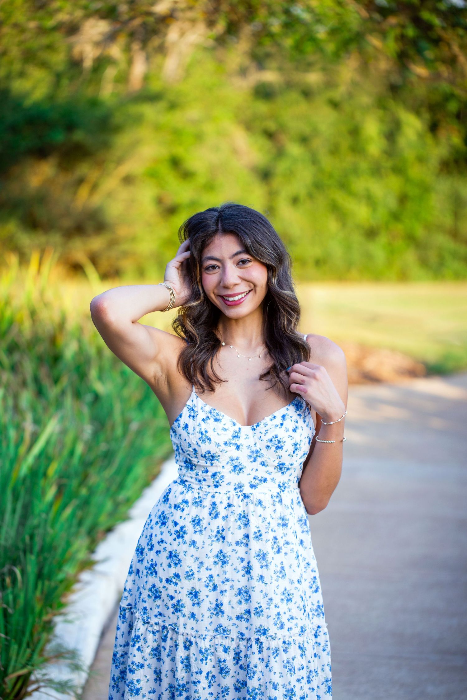 Texas A&M University graduate in a floral blue and white sundress is shown in an outdoor setting with greenery in the background.