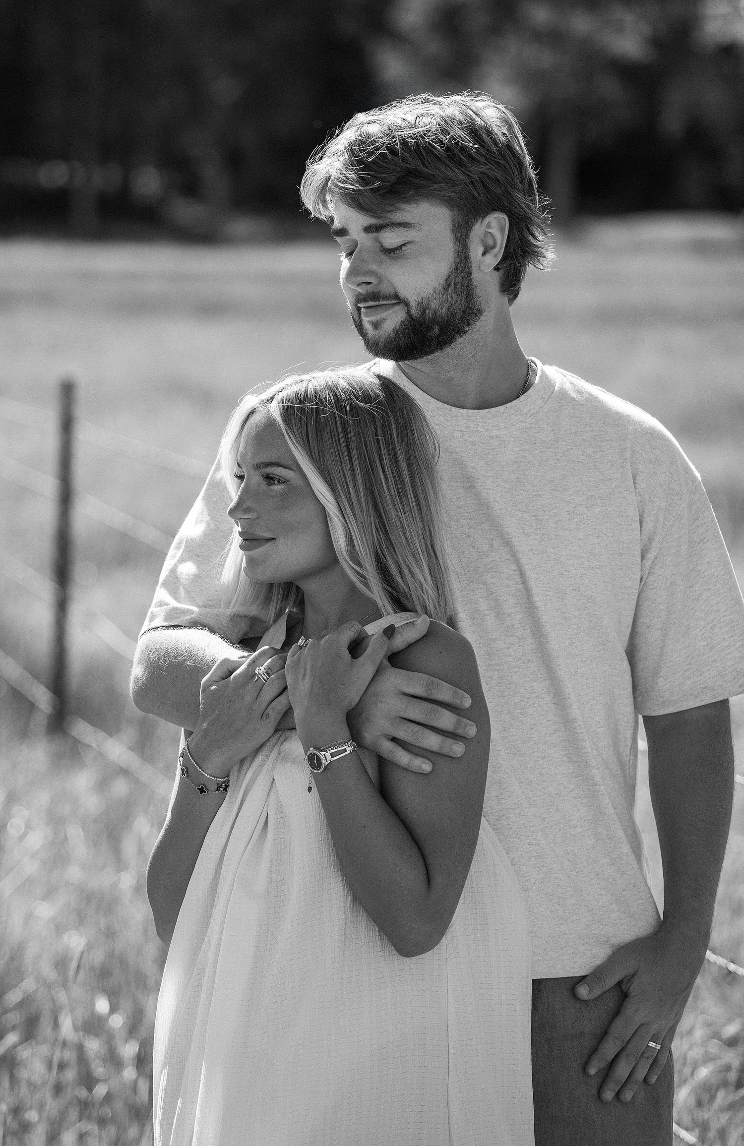Black and white photo of a couple embracing in a field, woman in white dress smiling, man in t-shirt holding her shoulders.